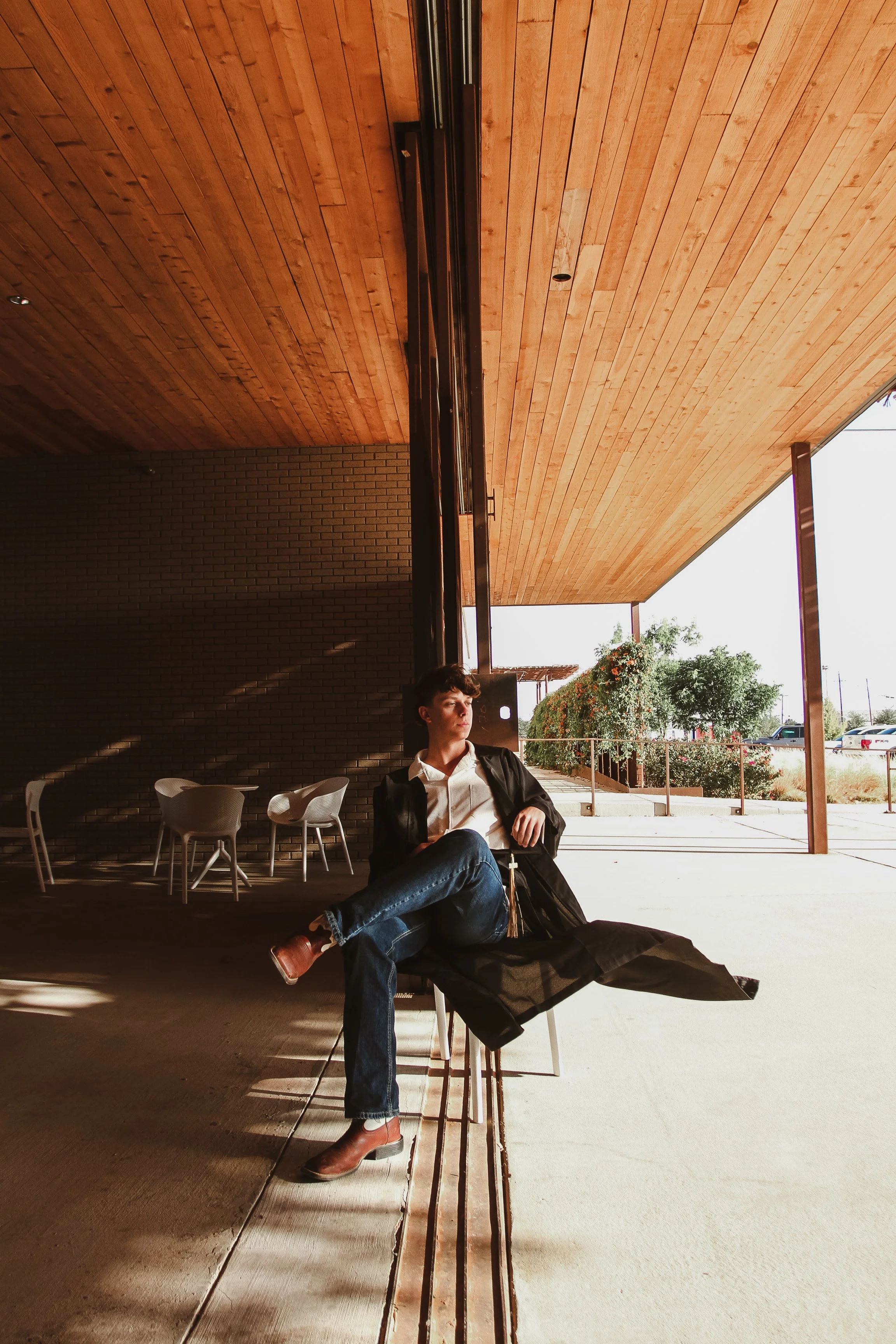A young man with dark hair, dressed in a white shirt, dark blazer, and jeans, sitting on a chair outside a building under a wooden roof, with sunlight casting shadows on the ground. There are white chairs and a brick wall behind him, and game or sports fields are visible in the background nearby.