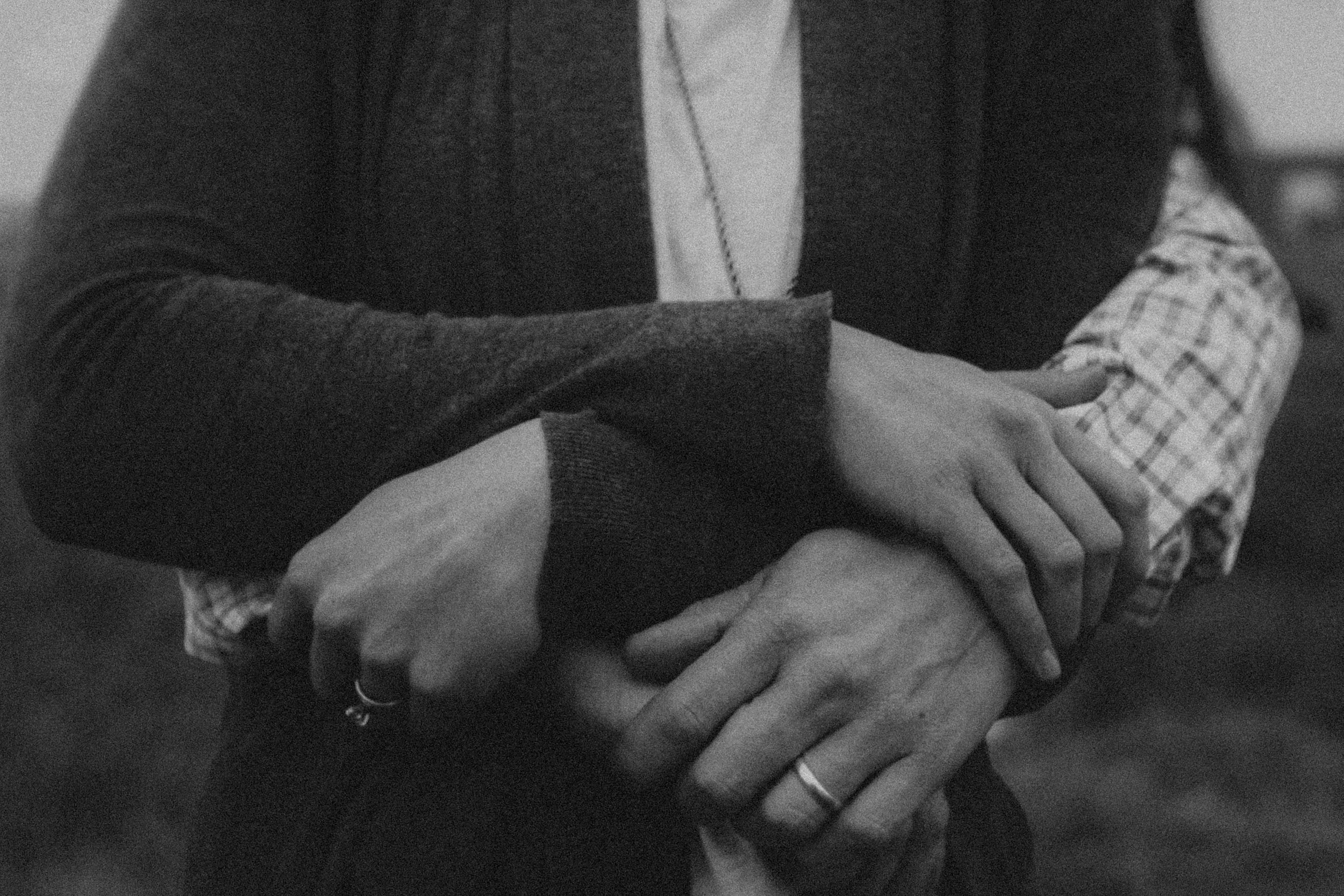 Close-up black and white photo of two people holding hands, one person's hand over the other's, with a background that is blurred.