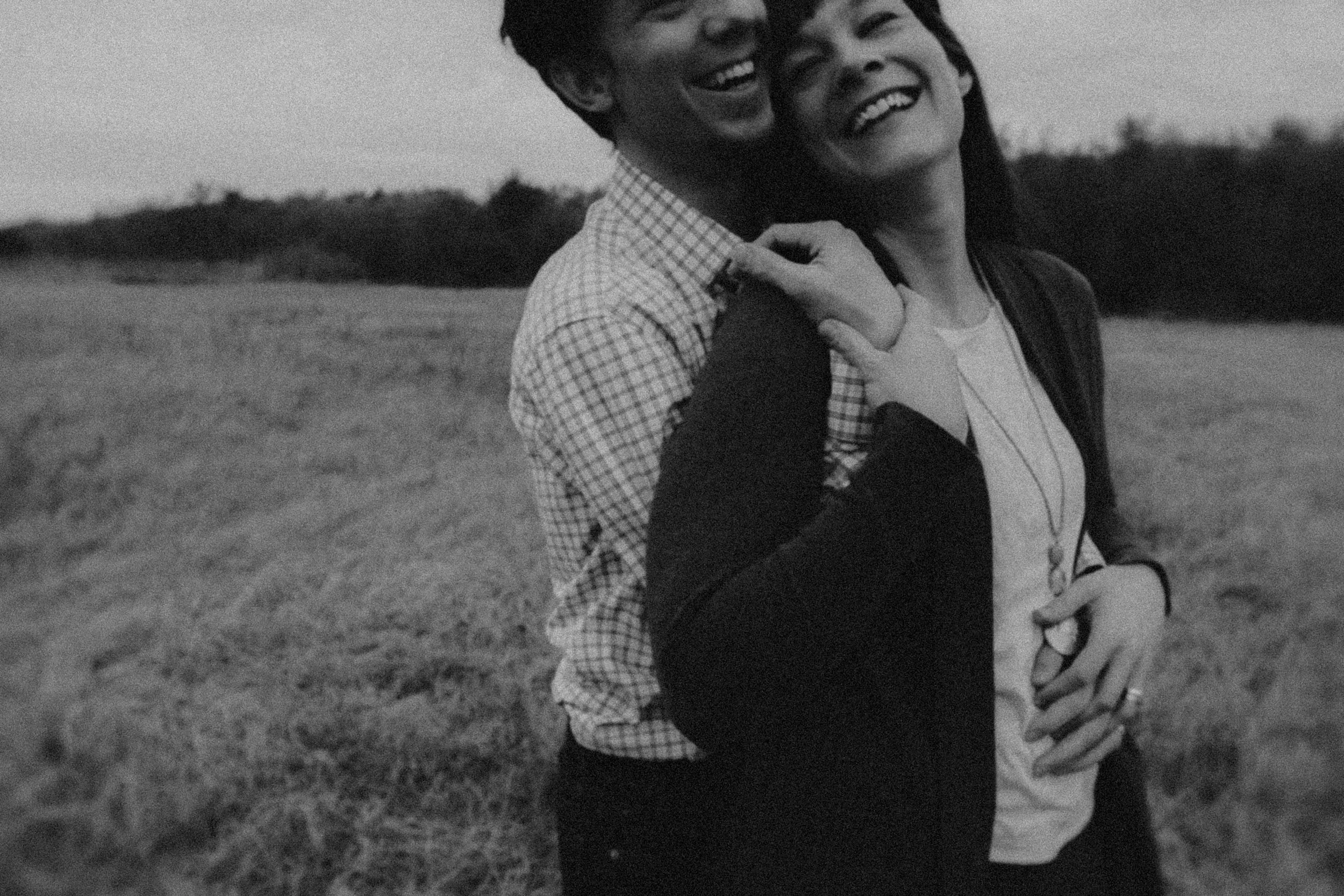 A black and white photo of a smiling young couple standing outdoors, close together, with blurred trees in the background.