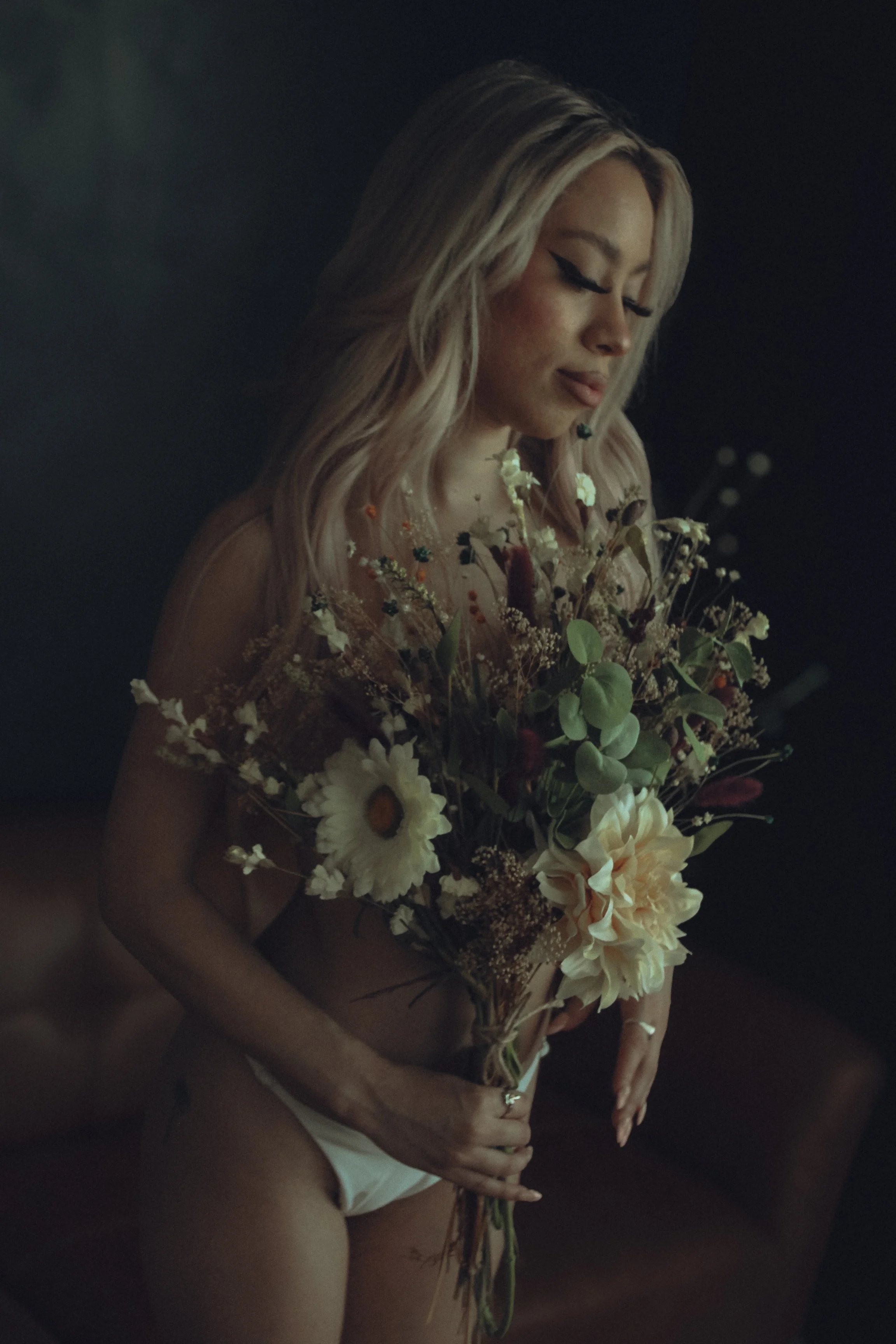 A woman with long, blonde hair holds a large bouquet of flowers, smiling softly with her eyes closed in a dark setting.