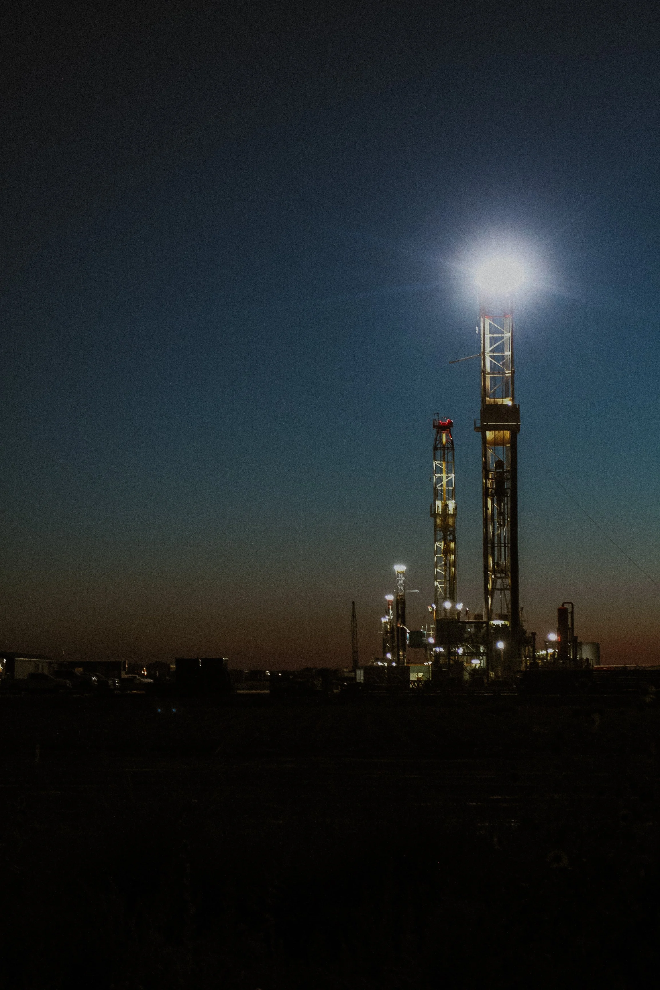 An oil drilling rig illuminated at night, with a dark sky and some parked vehicles in the foreground.