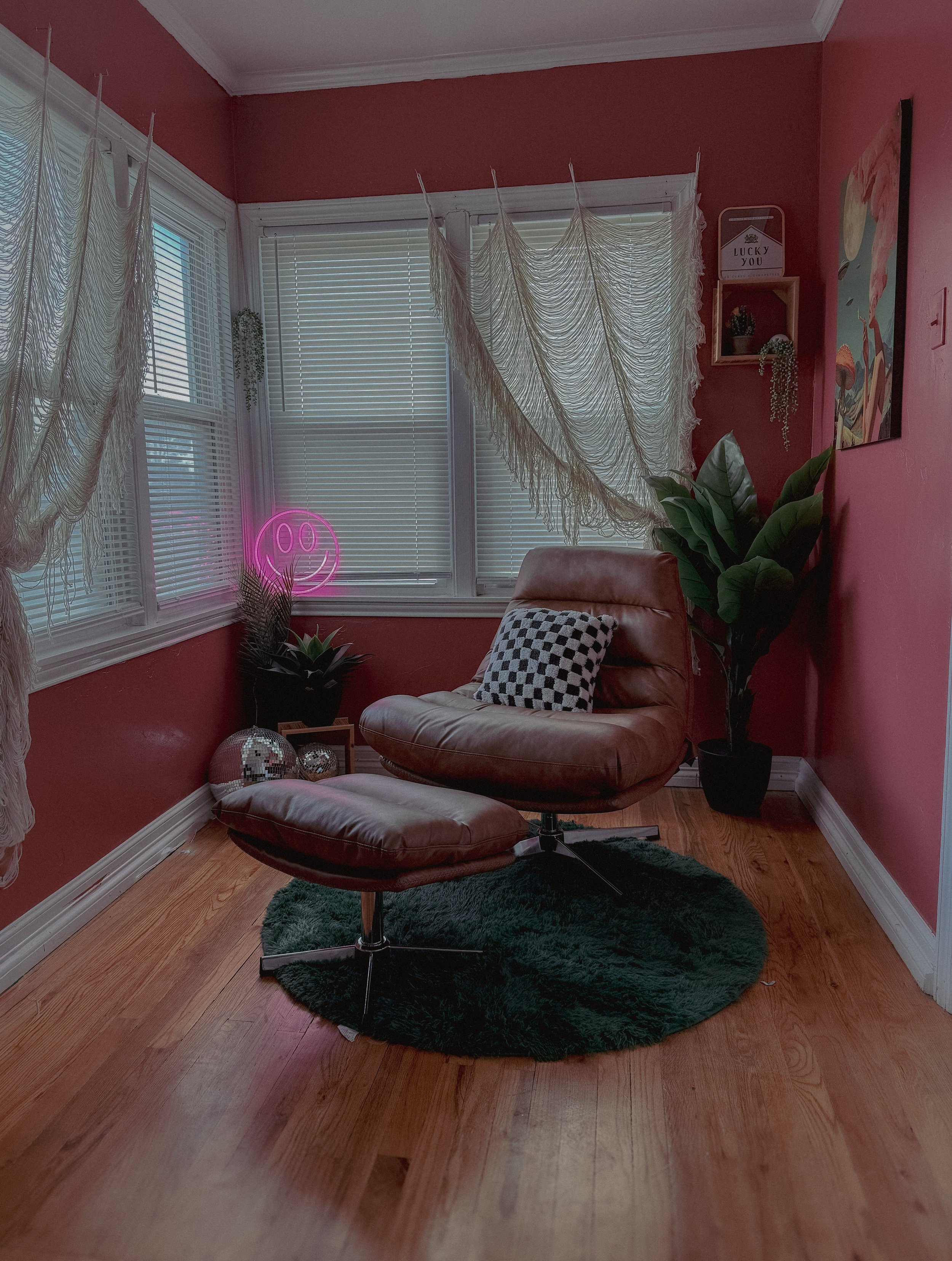 A cozy corner with pink walls, large windows with white blinds, and decorative elements including a tan leather lounge chair with a black-and-white checkered pillow, a dark green shaggy rug, plants, and wall art. A neon pink smiley face light adds ambiance.