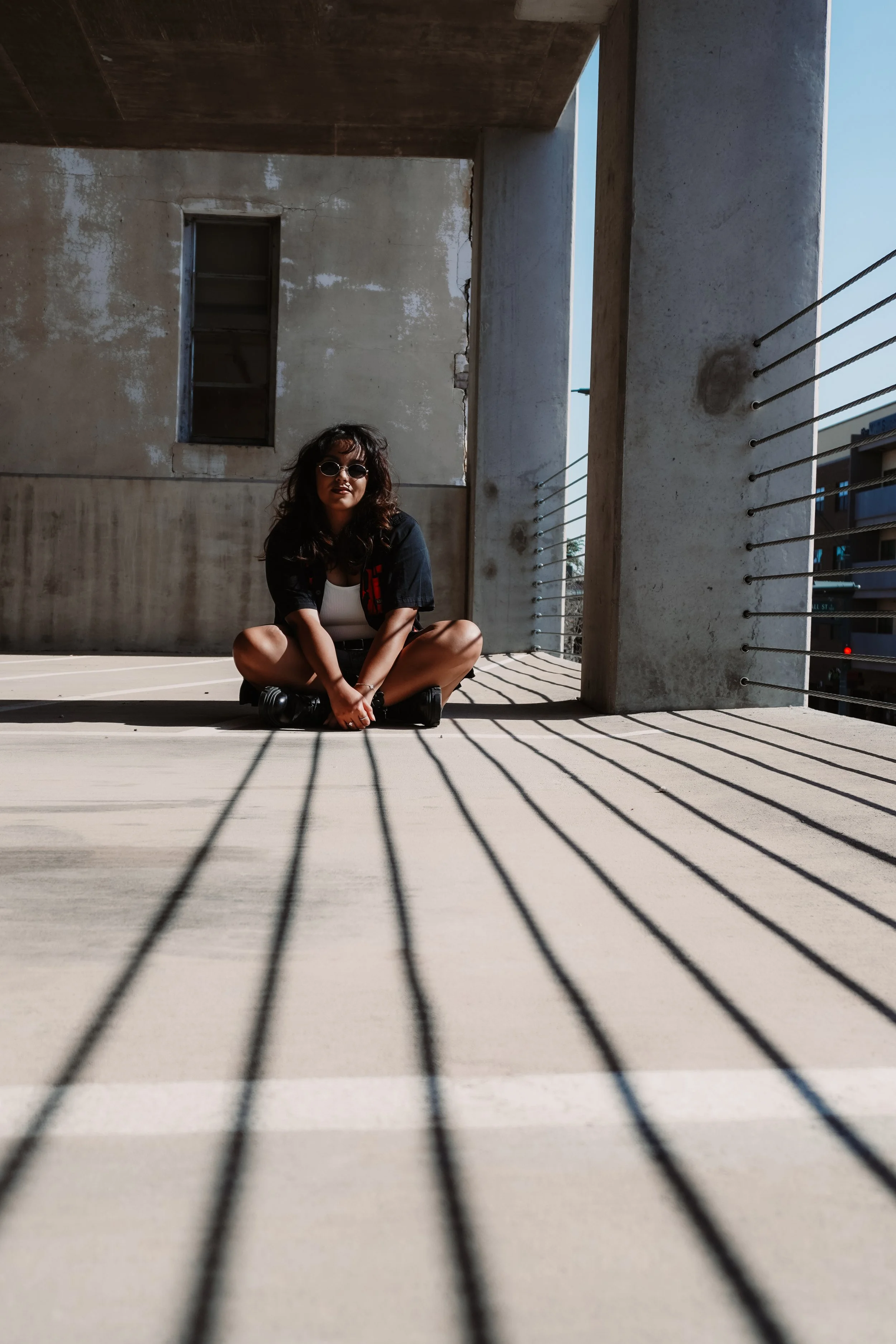 A woman with dark curly hair, wearing sunglasses, a white top, and black jacket, sits on the ground with her legs crossed in an outdoor urban space, with shadows cast by a railing on the ground and a concrete wall with a small, empty window behind her.
