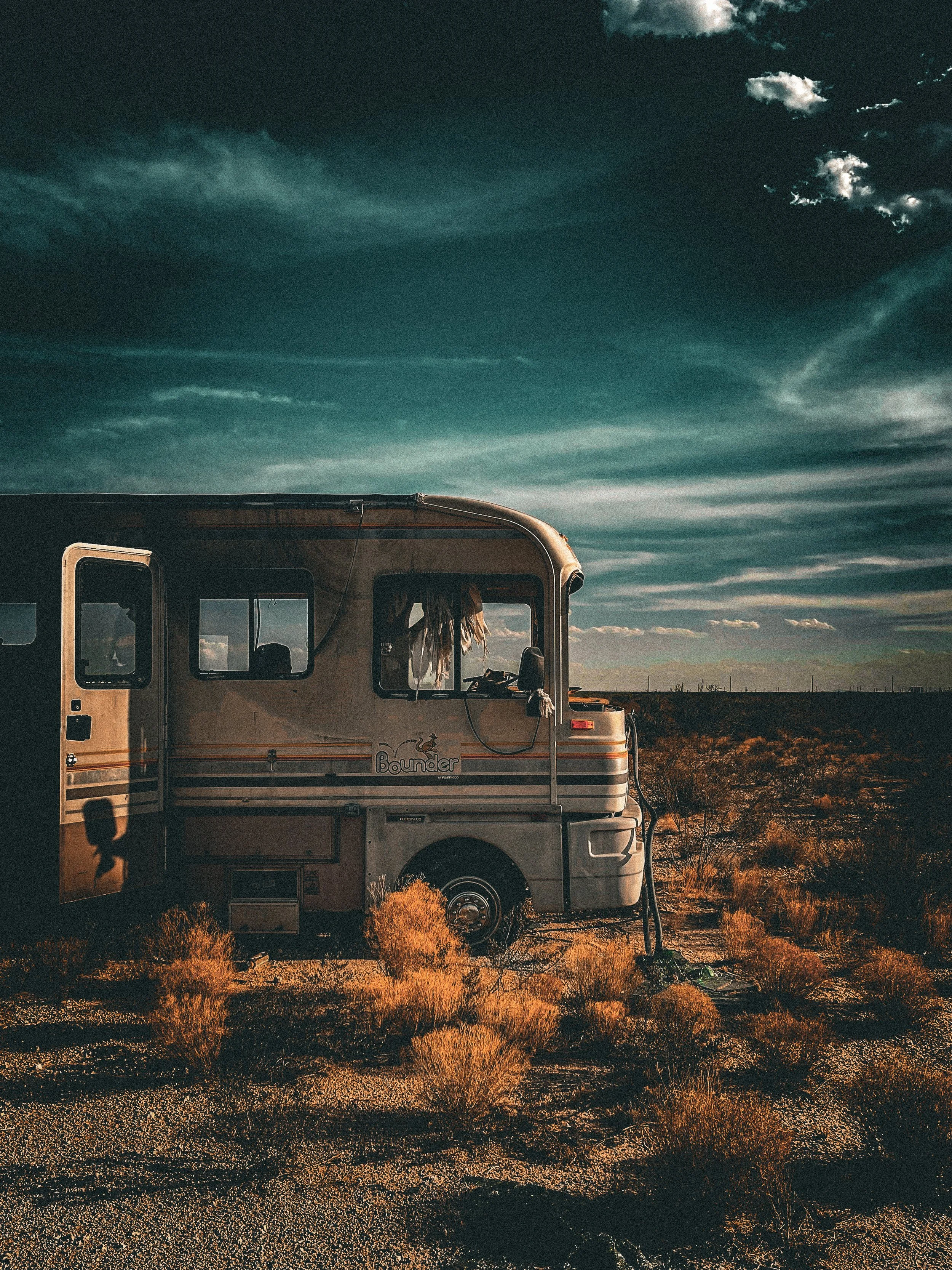 An orange and beige camper trailer labeled 'Bounder' parked in a desert landscape with dry shrubs, under a dramatic sky with dark clouds and some patches of sunlight.