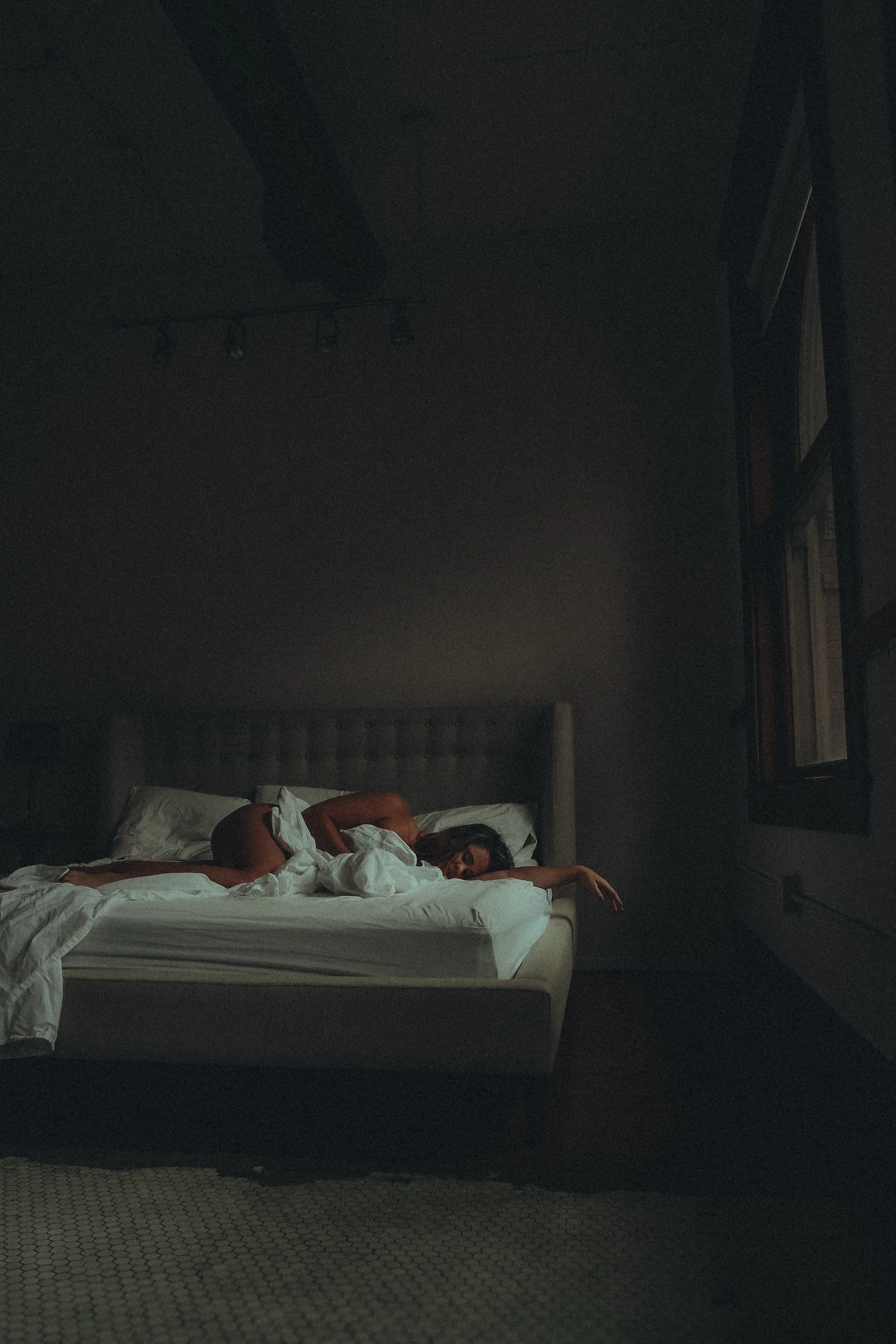 A woman lying on her side on a bed in a dimly lit bedroom, reaching her arm outward, with a head resting on pillows and a dark night sky visible through a window.