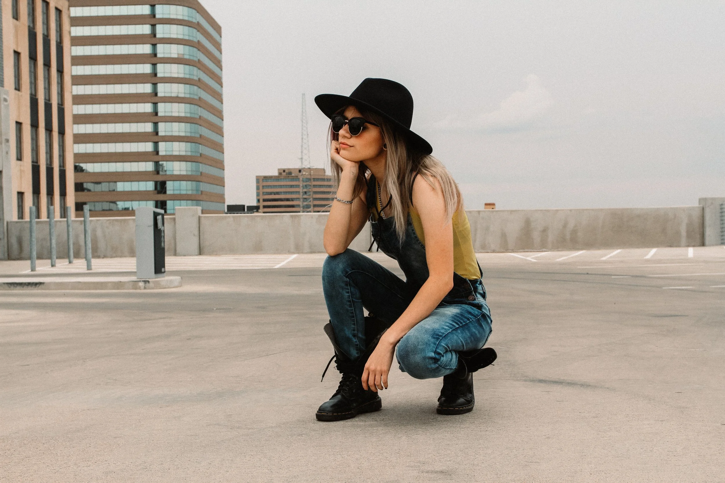 A woman with long hair wearing a black wide-brimmed hat, dark sunglasses, a yellow tank top, and black overalls crouches on the rooftop of a parking garage, with modern buildings in the background under a cloudy sky.