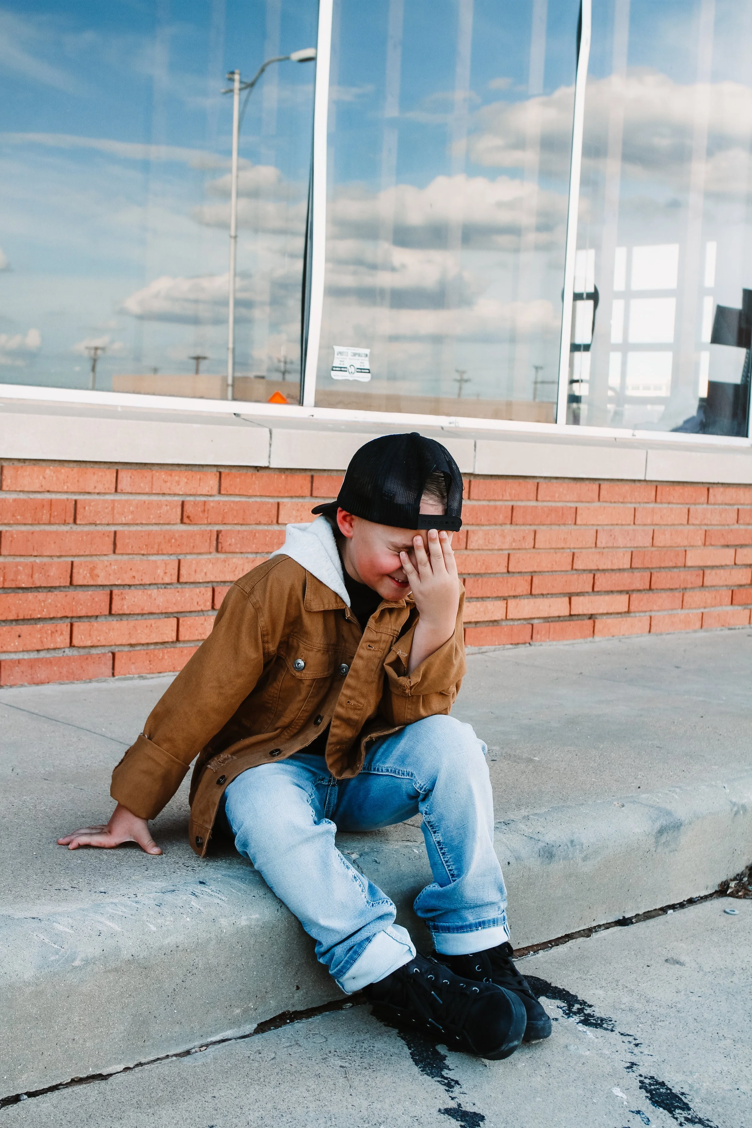 A young person wearing a jacket, hoodie, jeans, and sneakers sitting on the sidewalk with their hand covering their face, appearing distressed, in front of a brick building with large windows reflecting a blue sky and clouds.