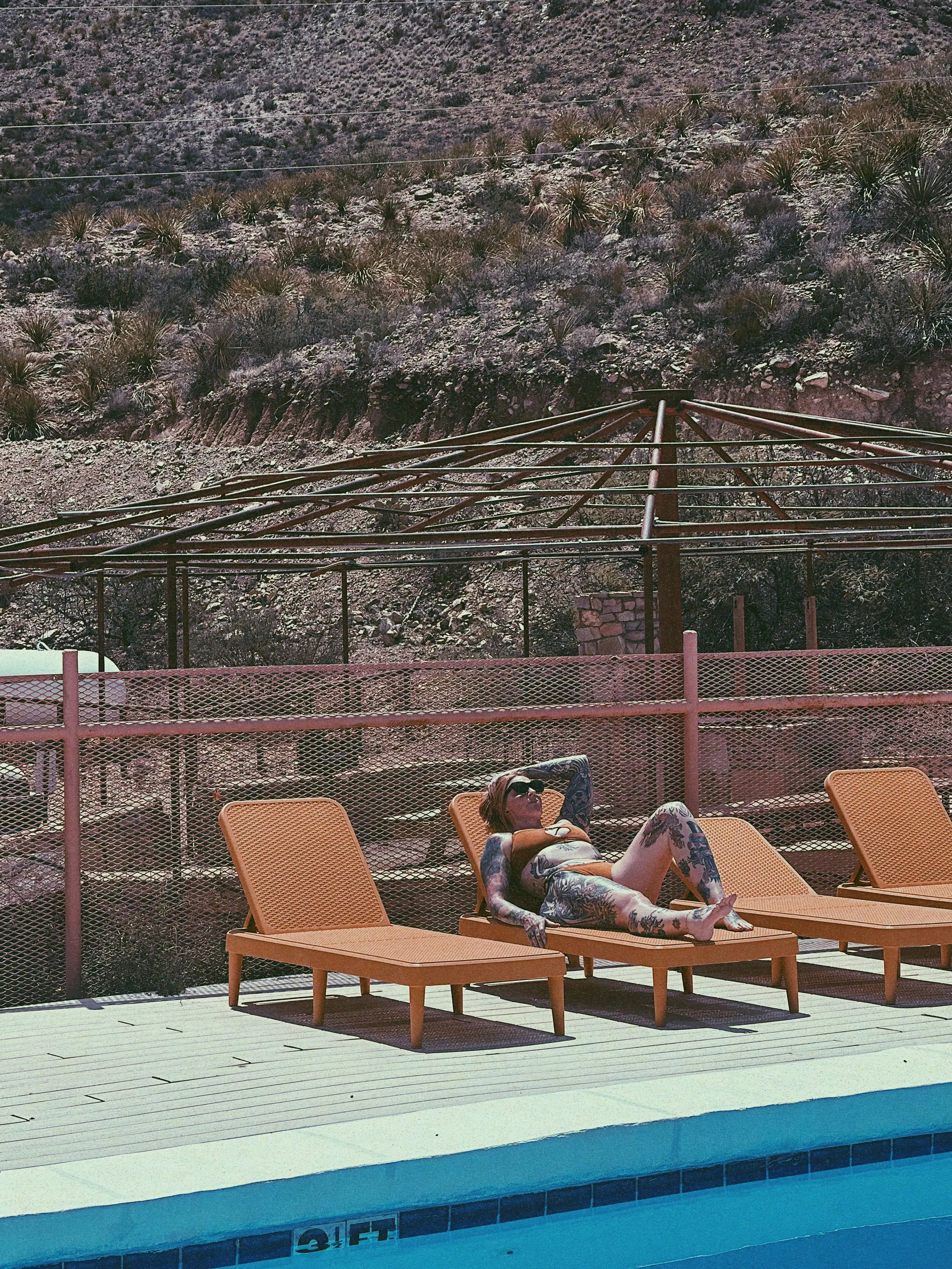 A person with tattoos relaxing on a lounge chair by a pool, with a mountain in the background.