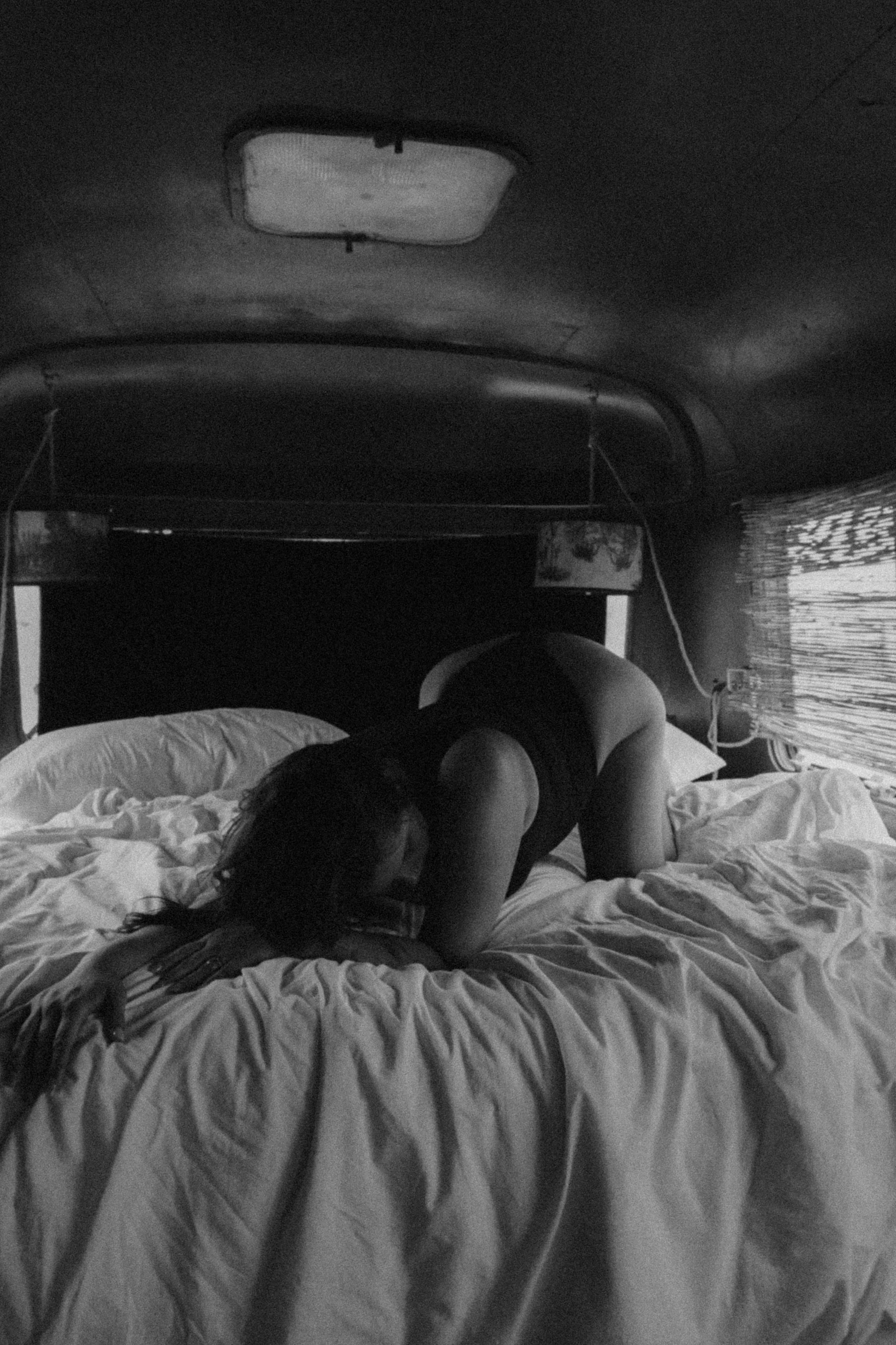 A person with long dark hair is lying face down on a bed with white sheets, in a room with dark walls and a window with blinds. The person appears to be resting or sleeping.