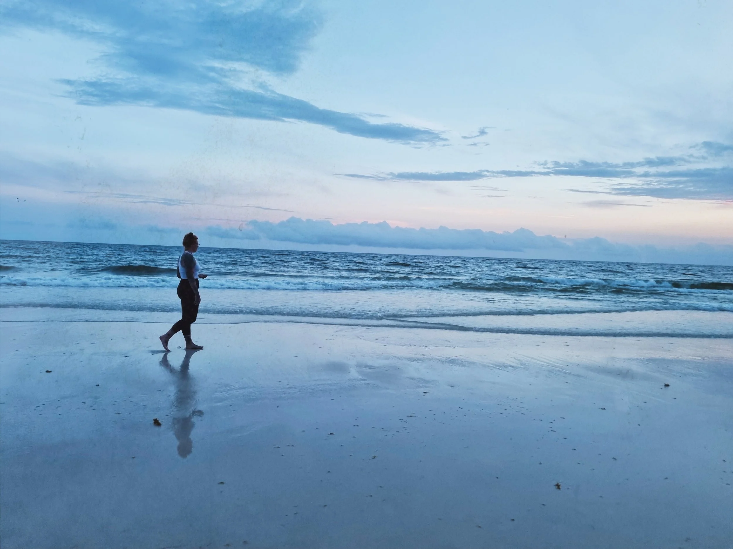A woman walking on the wet sandy beach near the shoreline during sunset. The sky is mostly cloudy with hints of pink and blue, reflecting on the water and wet sand.