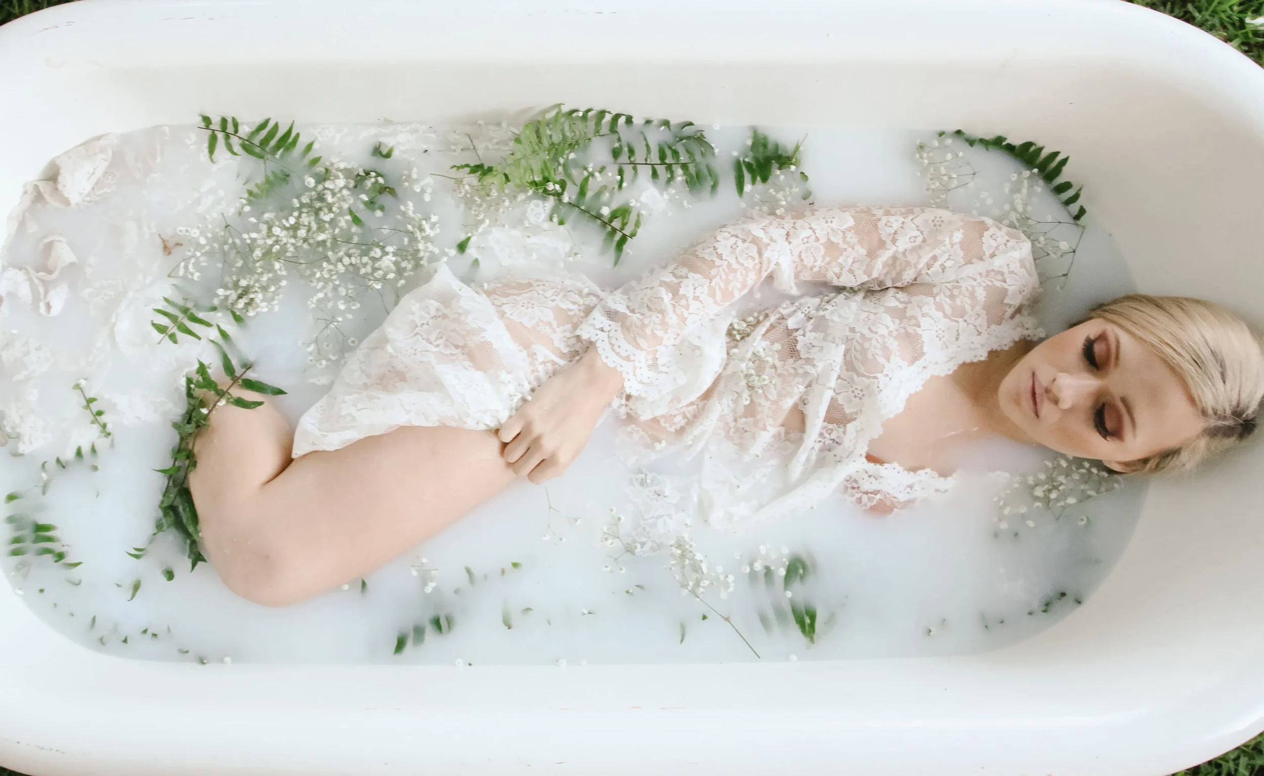 A woman lying in a bathtub filled with a white milky bath surrounded by green fern leaves and white baby's breath flowers, wearing a lace robe and smiling with eyes closed.