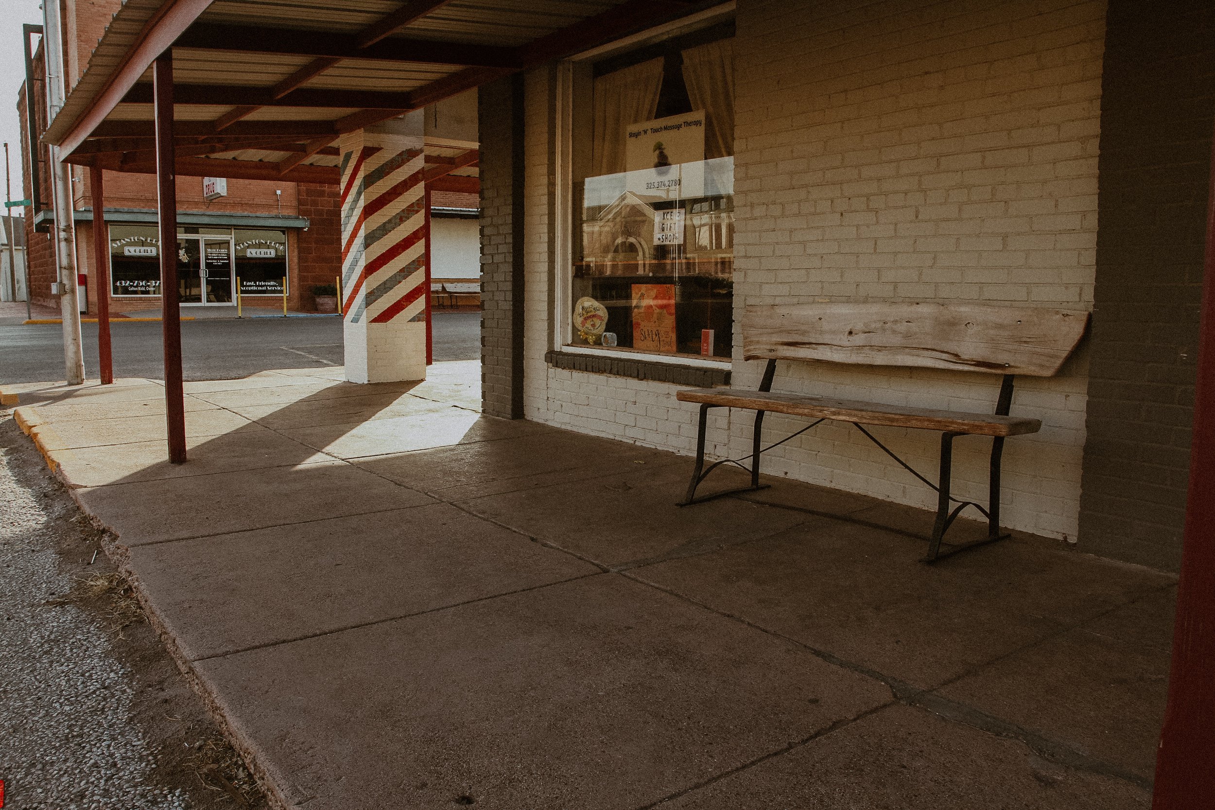 Empty sidewalk outside a brick building with a large display window, a wooden bench, and a barber pole painted red, white, and blue.