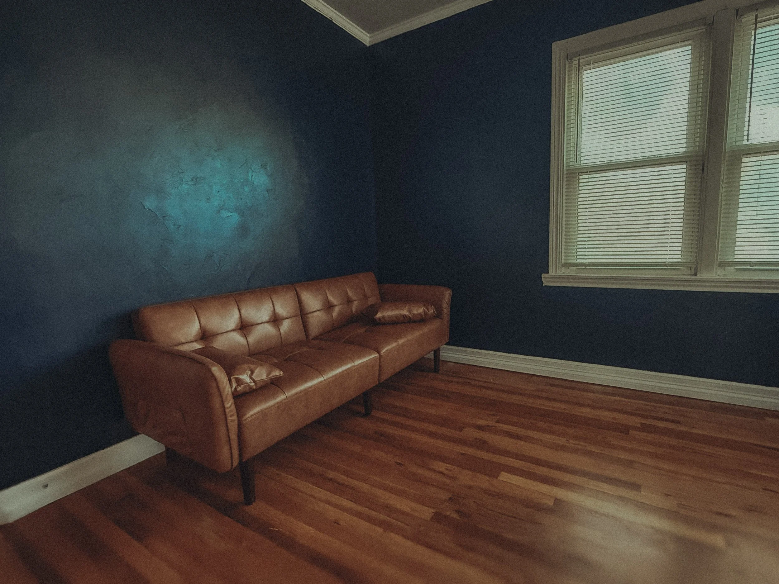 A room with dark blue walls, a wooden floor, a large window with plantation shutters, a brown leather couch, and a small cushion on it.