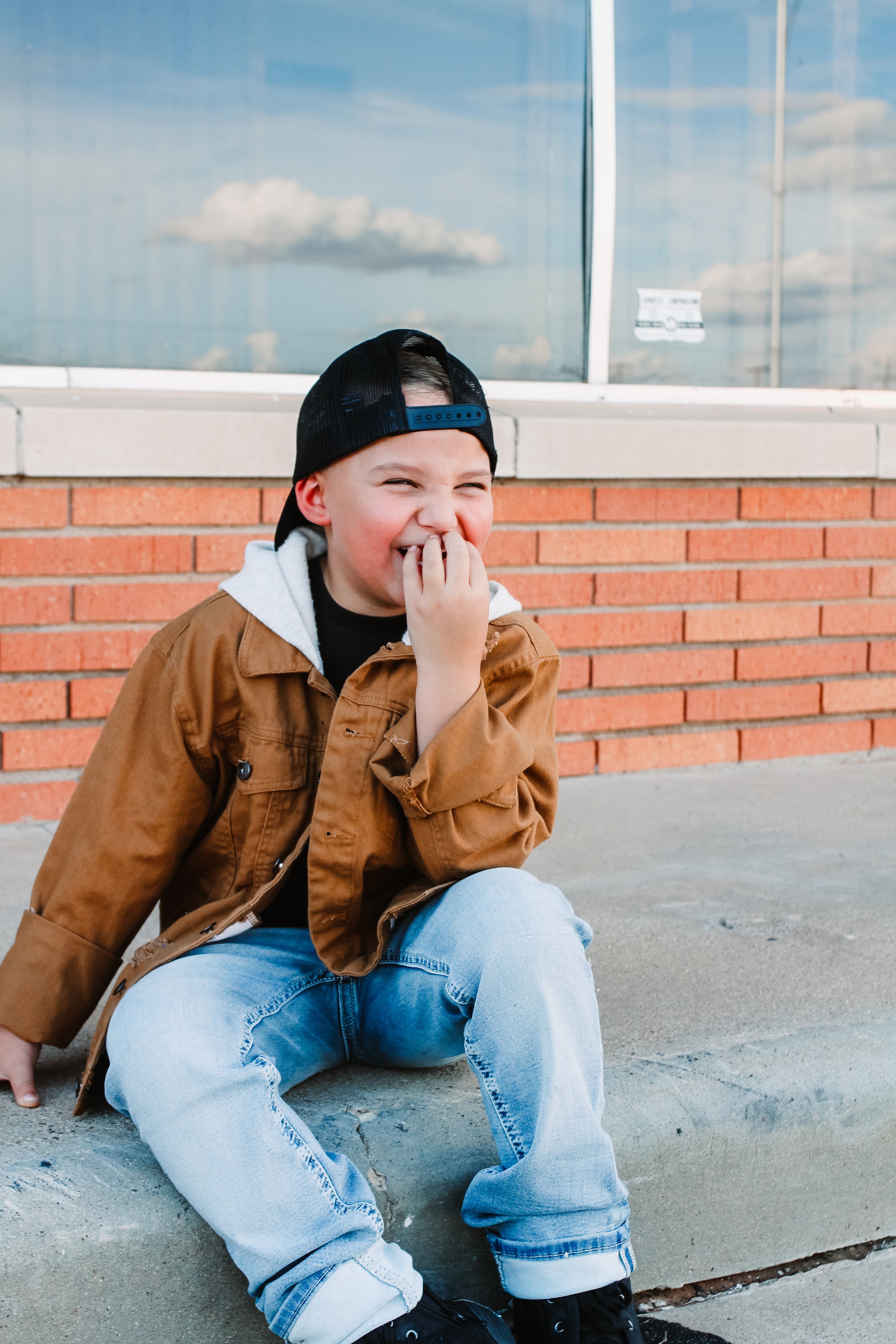 A young boy is sitting on the sidewalk laughing, wearing a black cap backwards, a brown jacket with a gray hoodie underneath, light blue jeans, and black shoes. He is covering his mouth with his left hand and looking to his left, with a brick wall and large glass windows reflecting the sky and clouds in the background.