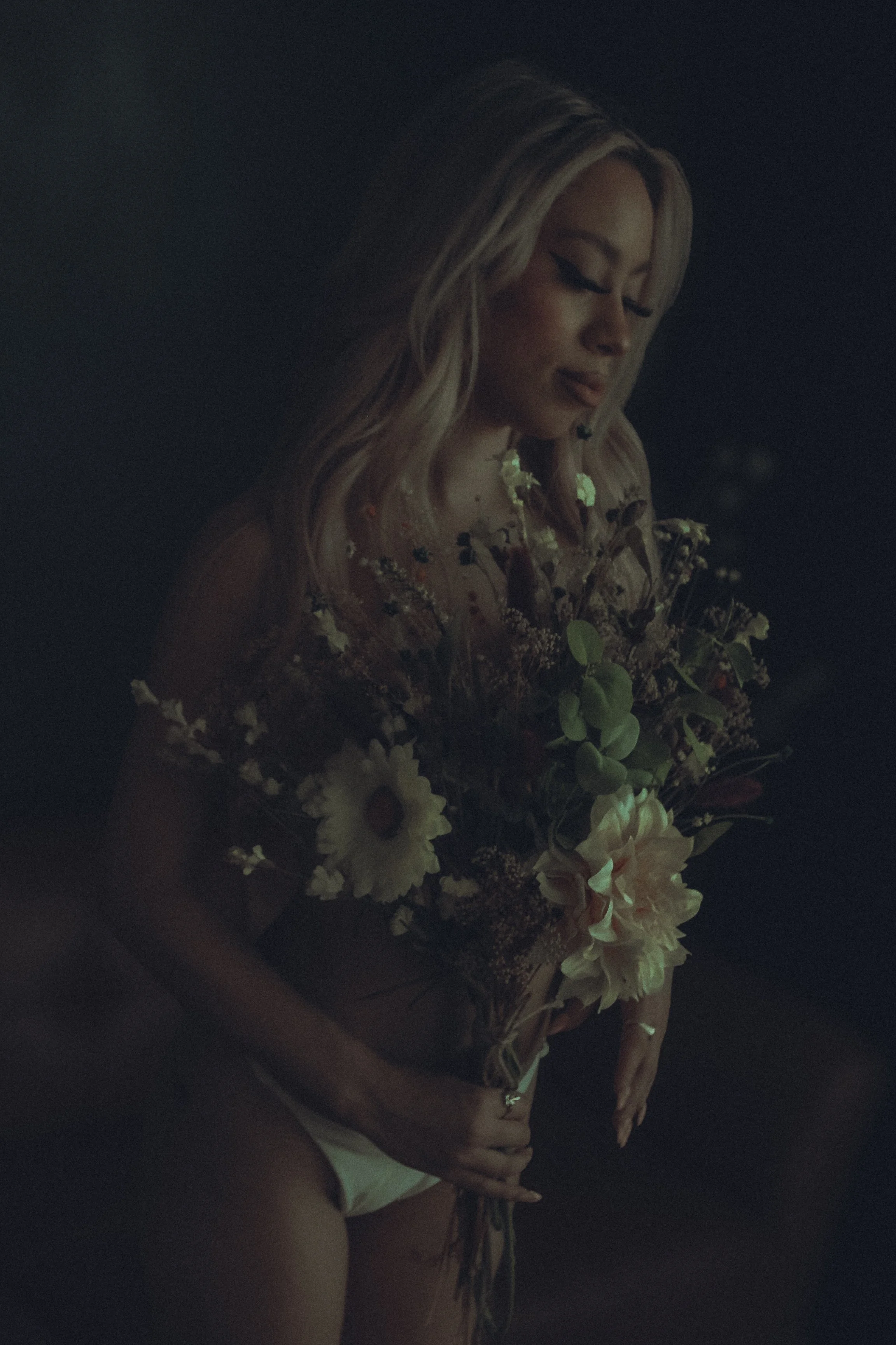 A woman with long blonde hair holding a bunch of flowers in a dark setting.