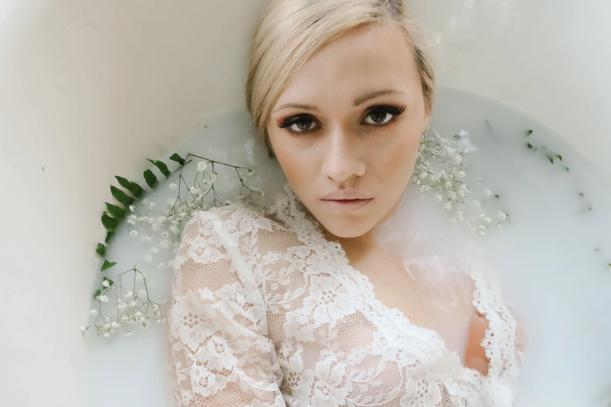 A woman with blonde hair taking a partially submerged selfie in a white bathtub filled with water, surrounded by small white flowers and green leaves.
