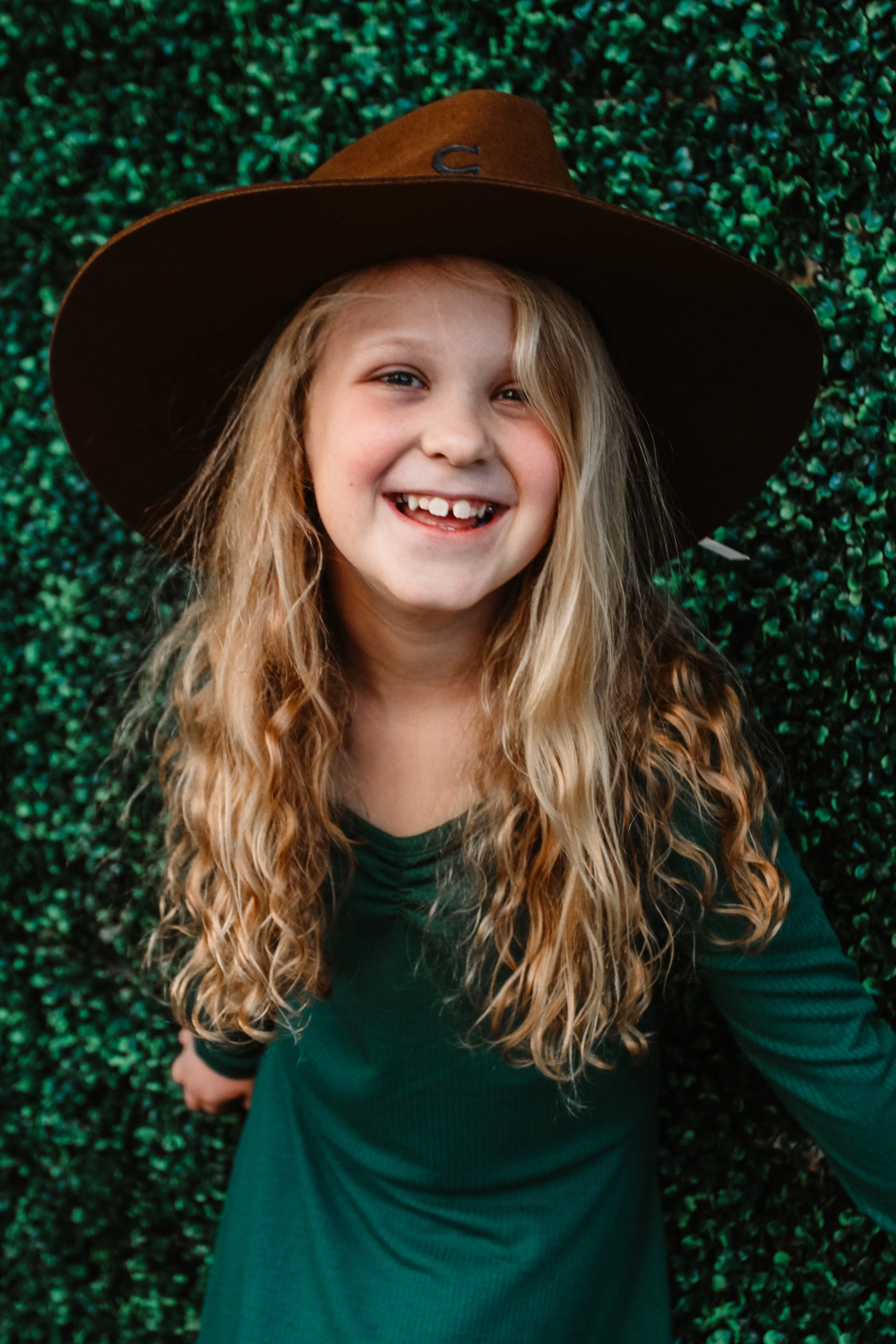 A young girl with long blonde curly hair, wearing a large brown cowboy hat with a small embroidered letter C, smiling widely with missing teeth, standing in front of a green leafy background.