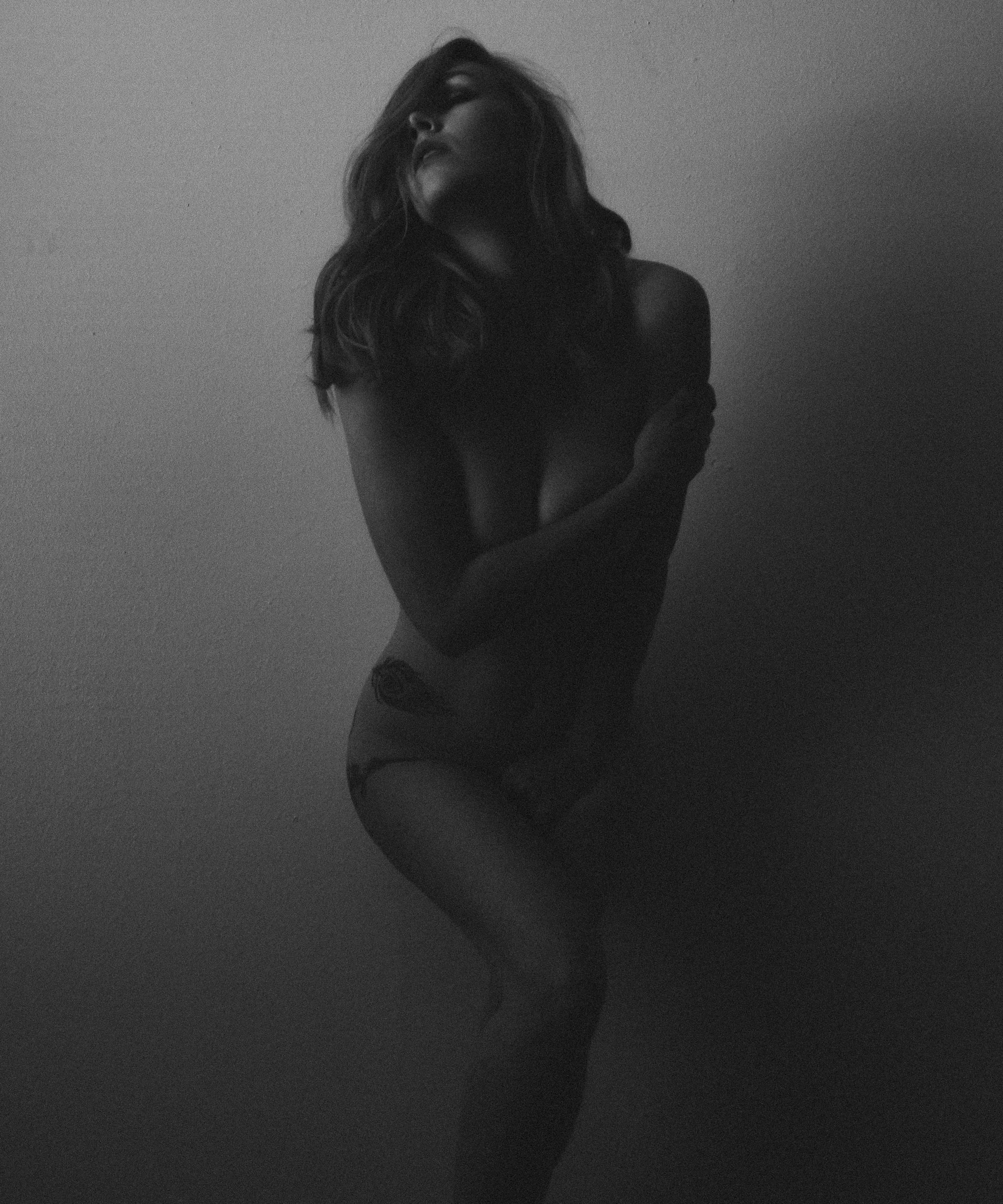 Black and white photograph of a woman with long hair, her eyes closed, her head tilted back, and her arms crossed over her chest, standing against a plain wall.