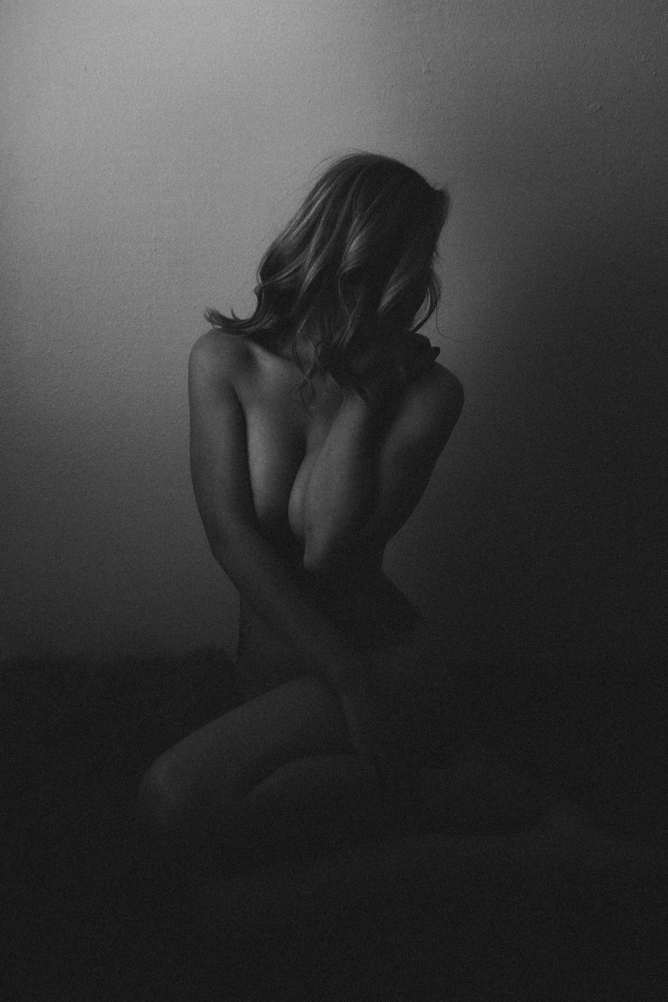 A black and white photo of a woman with long wavy hair, sitting with her head bowed, arms crossed over her chest, and her face partially obscured.
