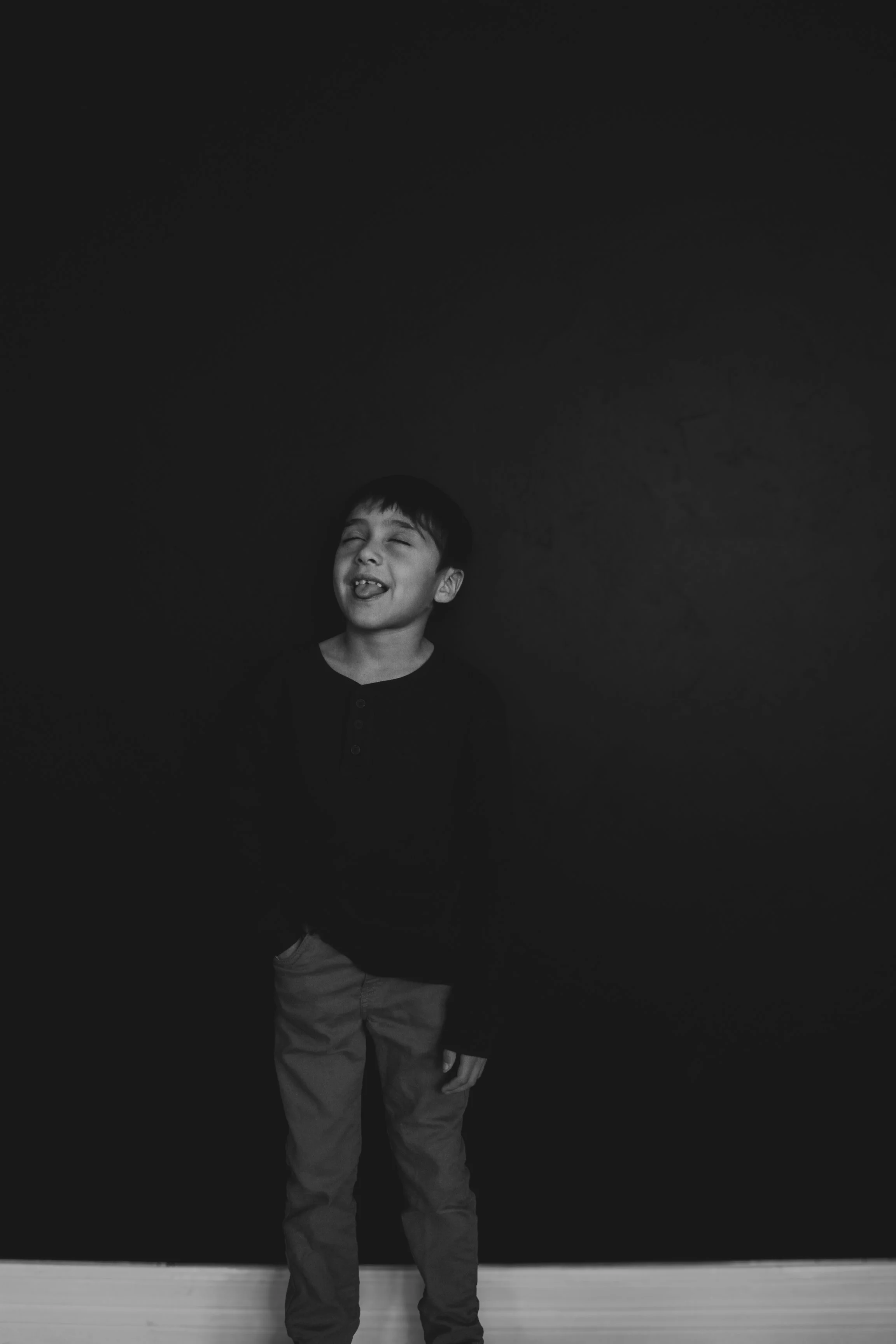 A young boy standing in front of a black wall, smiling and sticking out his tongue.