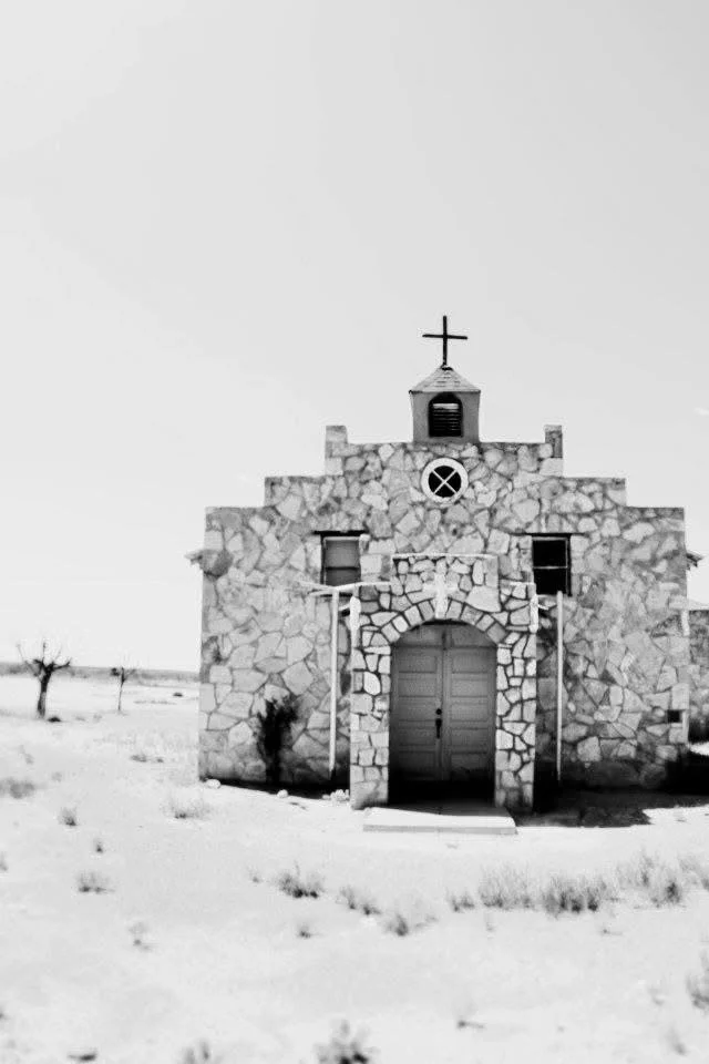 Stone church with a cross on top, set in a snowy landscape with a few barren trees.