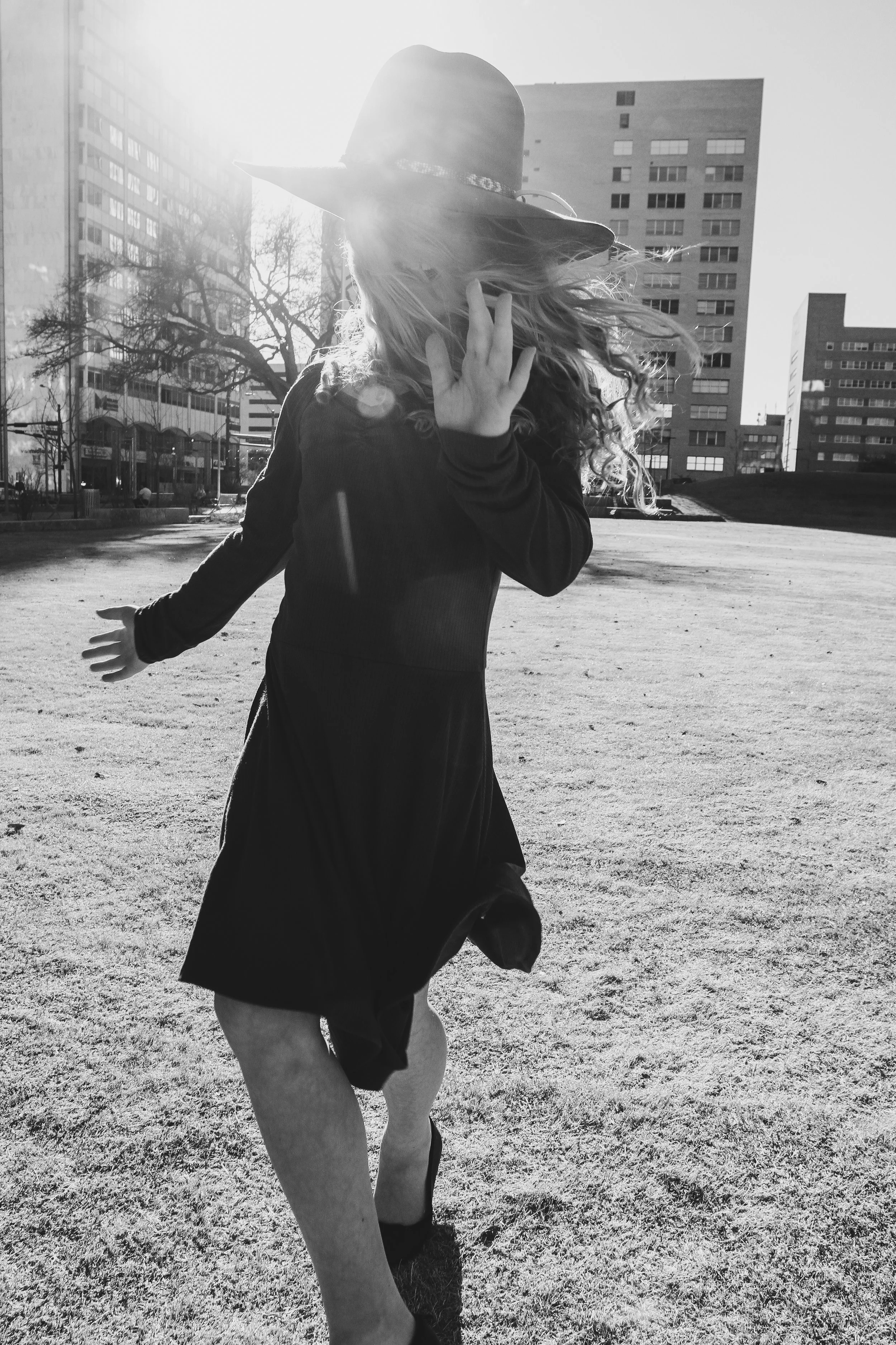 A woman wearing a hat and a dress running in a park with tall buildings in the background, captured in black and white.