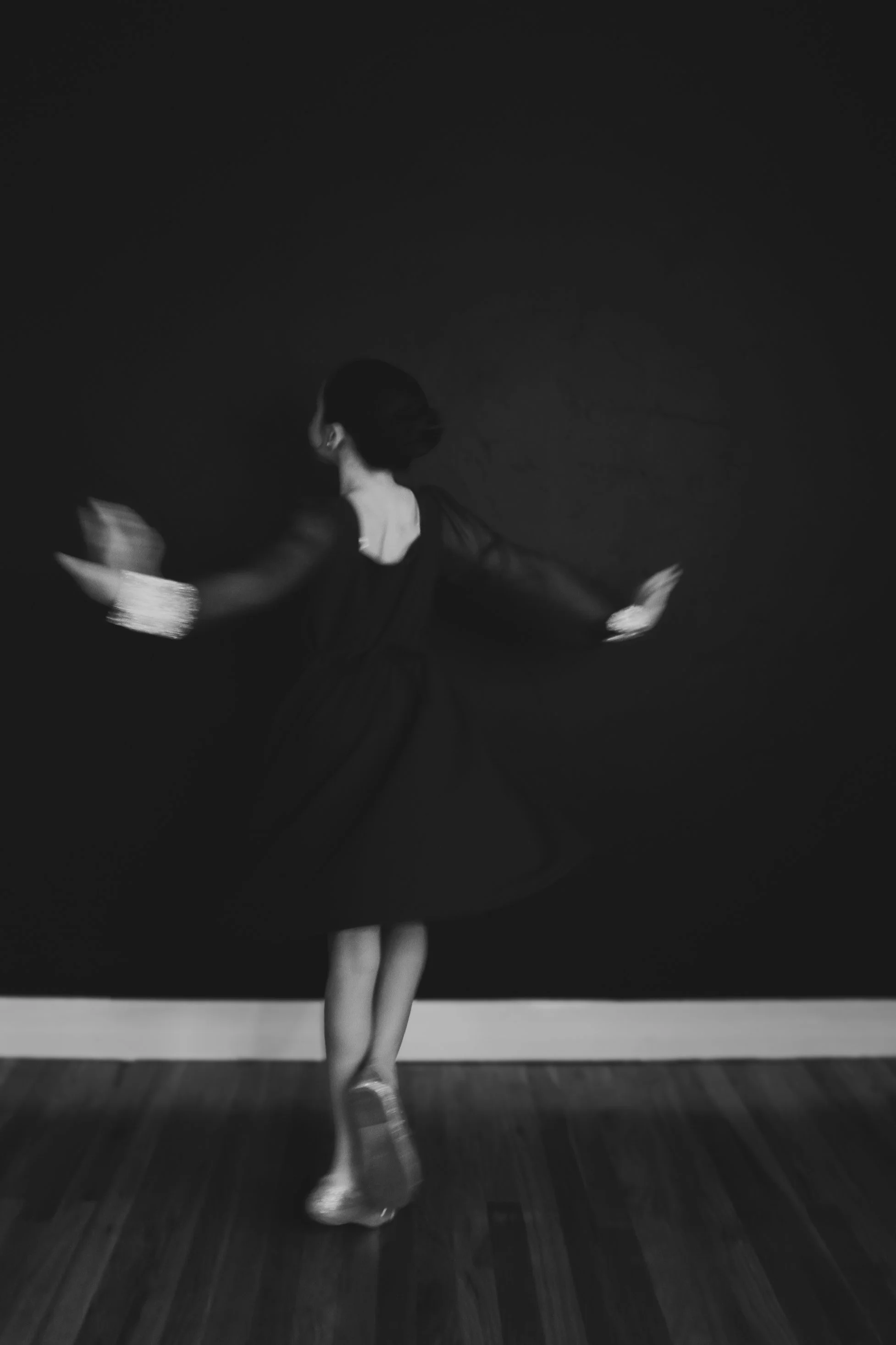 A young girl in a dance costume performing a ballet move on stage, captured in motion with her arms raised and one leg lifted, against a dark background.