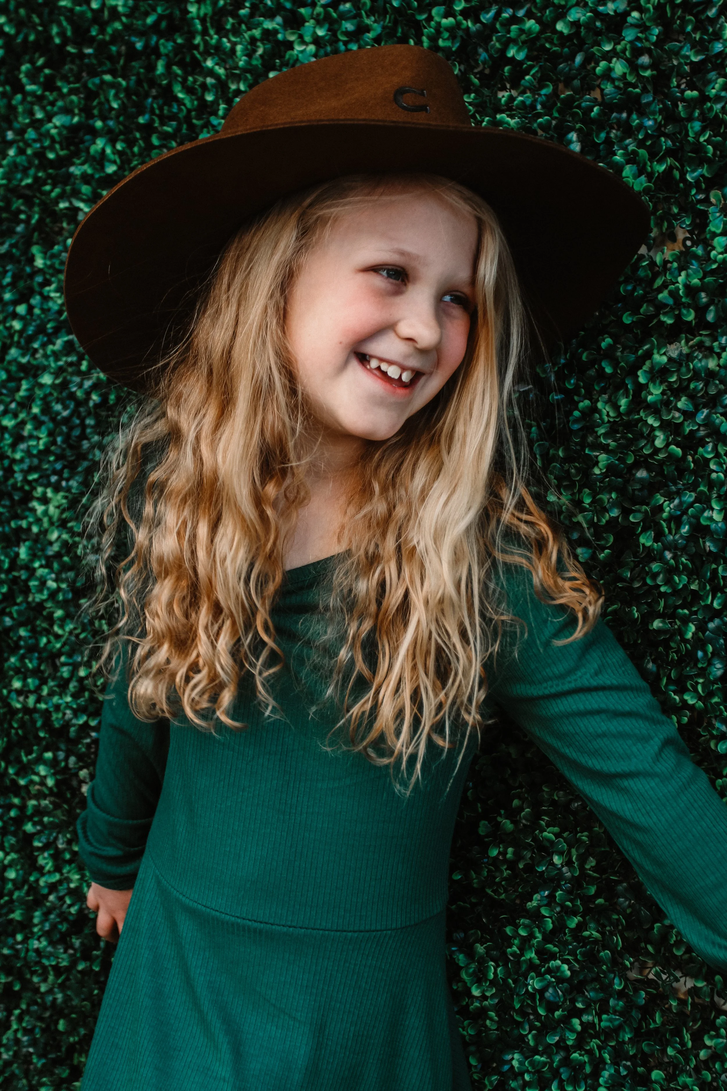 A young girl with long, wavy blonde hair wearing a dark green long-sleeve shirt and a large brown wide-brimmed hat, smiling and looking to her left, standing against a dark green, leafy background.