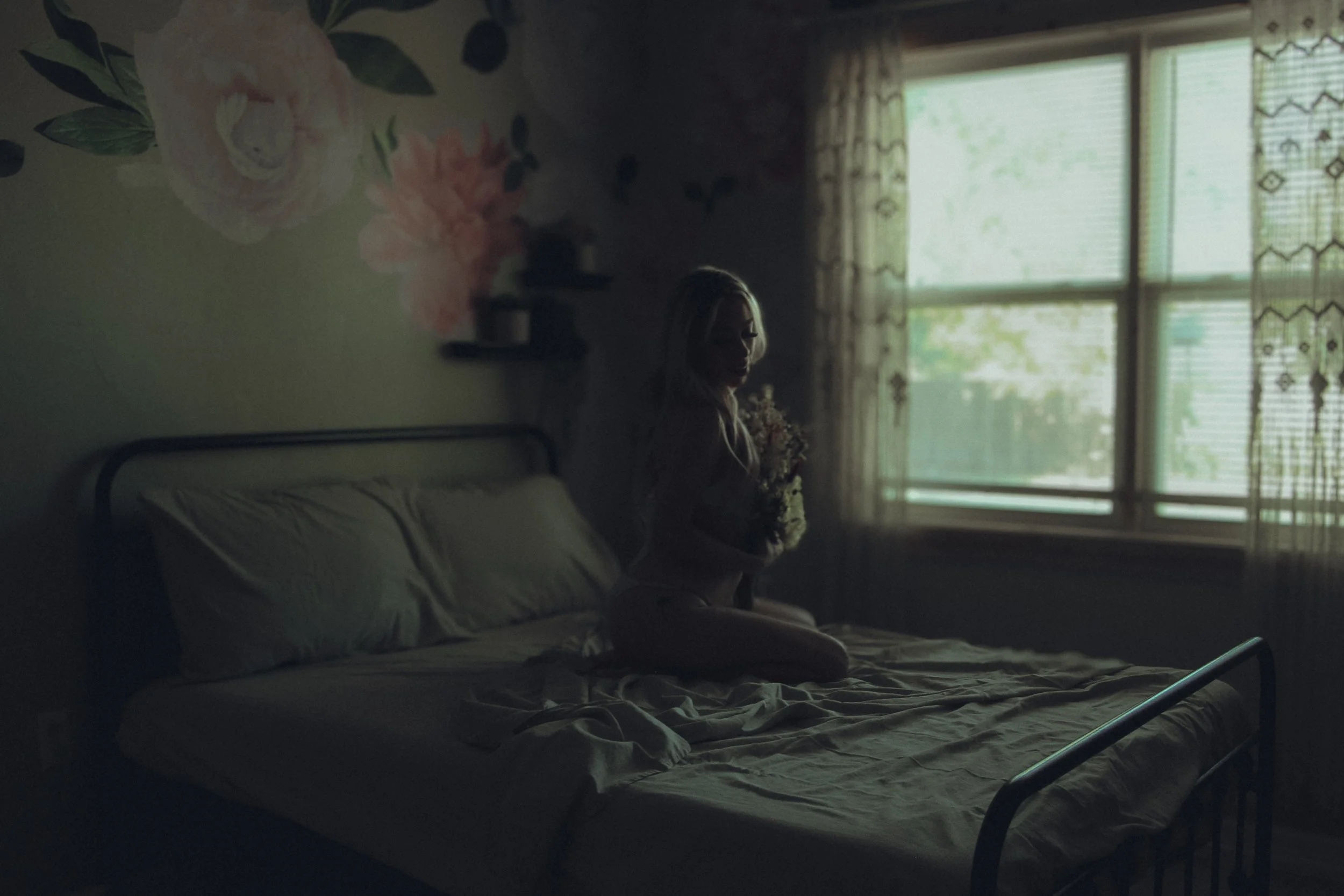 A woman with blonde hair and glasses sitting on a hospital bed, holding a bouquet of flowers. The room is dimly lit with natural light coming through the window, which has patterned curtains. The wall has floral wallpaper and a shelf with small objec