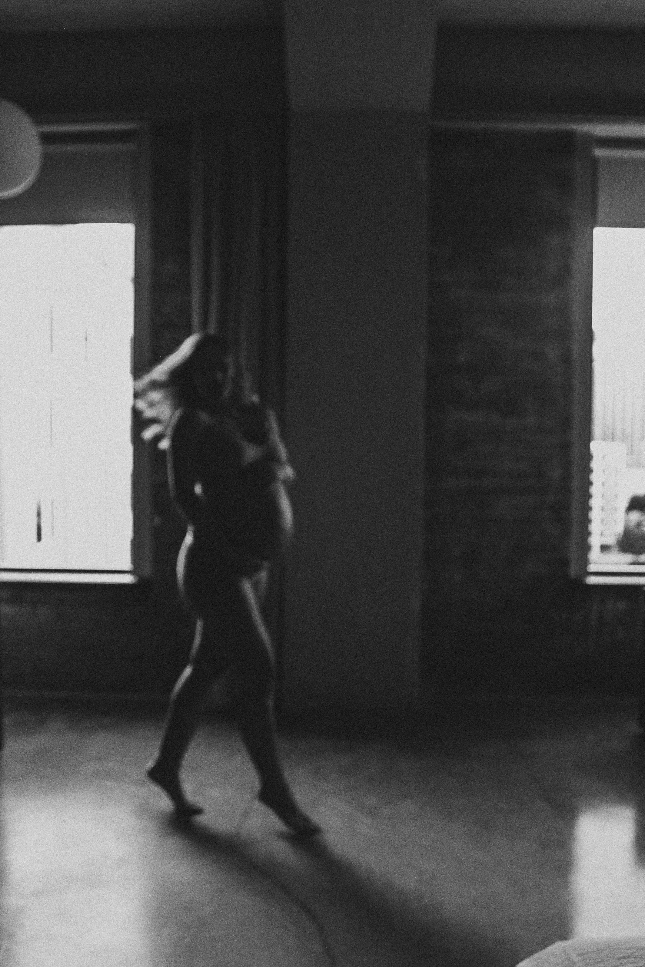 A woman in black attire walking barefoot in a room with what appears to be a large mirror, with reflections of windows and blinds.