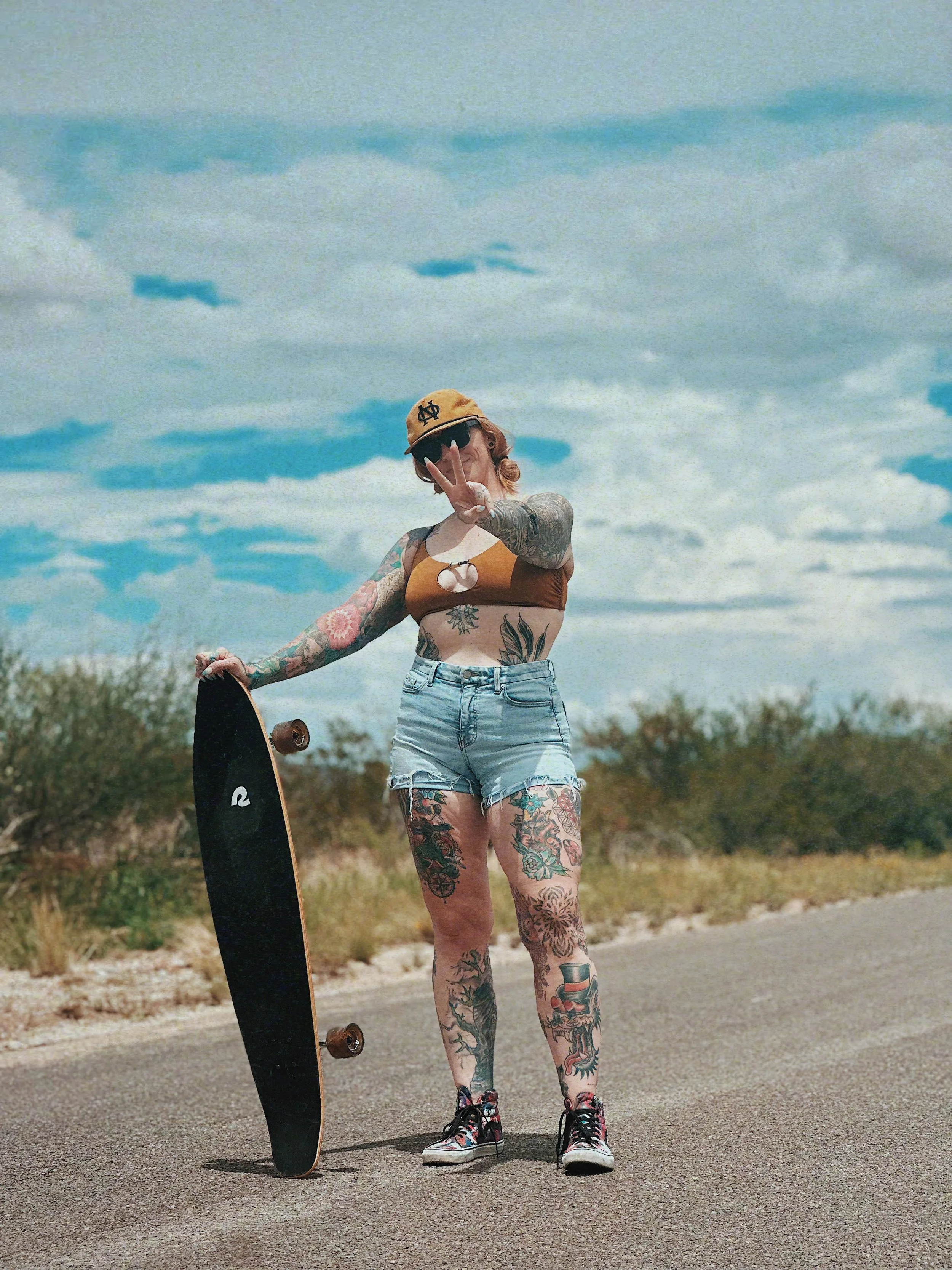 A tattooed woman wearing a tan baseball cap, sunglasses, a brown crop top, distressed denim shorts, and high-top sneakers stands on a road holding a skateboard. She is making a peace sign with her right hand, and the sky is partly cloudy with trees in the background.