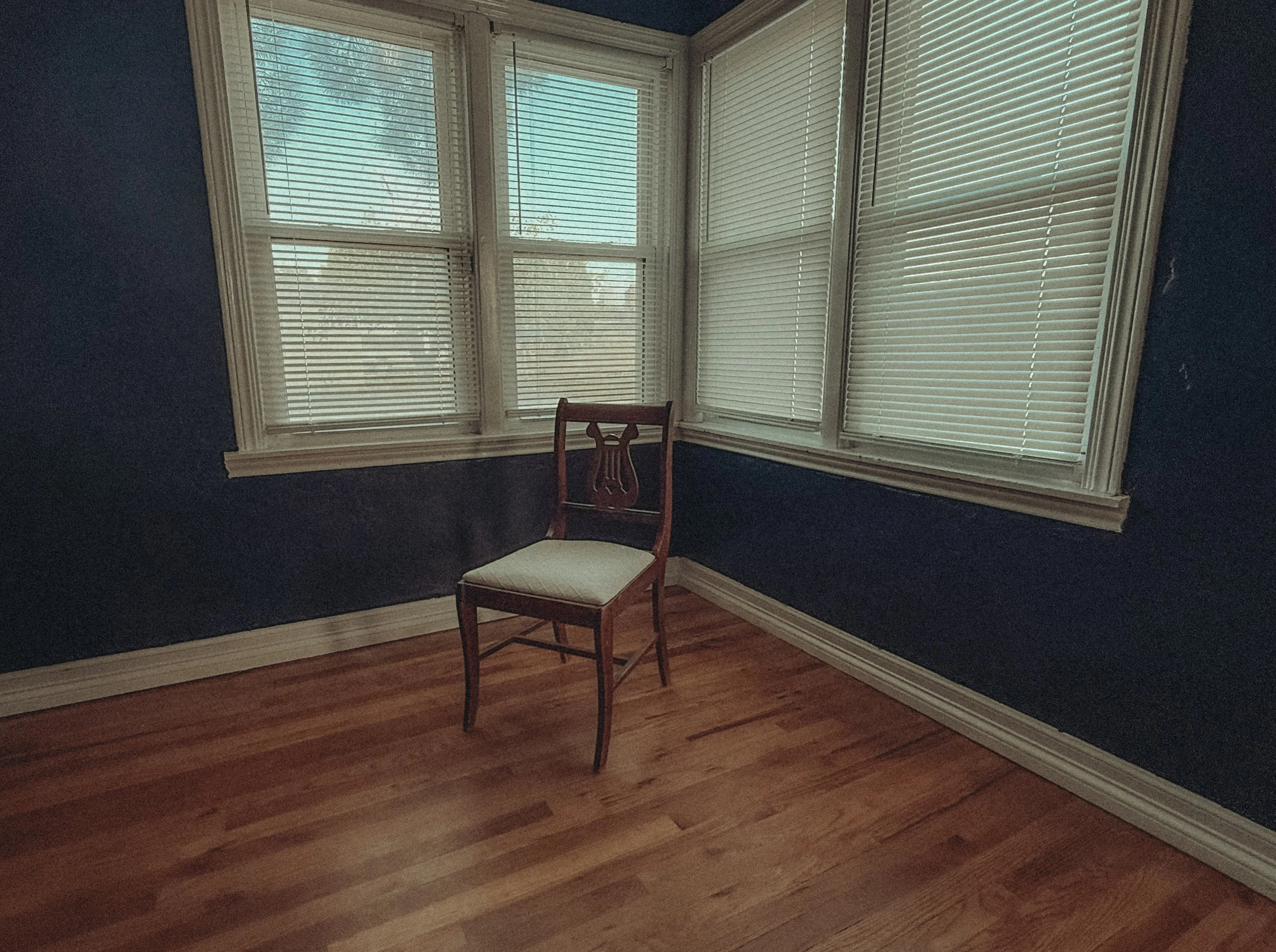 A corner of a room with dark blue walls, a wooden floor, and white window blinds covering large windows; there's a wooden chair with a white cushion near the corner.