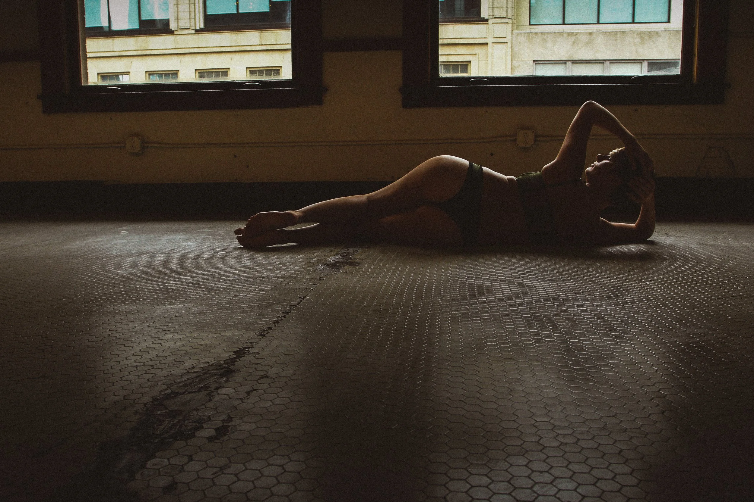A woman lying on the floor in a dimly lit room, with two open windows showing building facades outside, wearing dark underwear and a top, with one arm behind her head and the other resting on the floor.