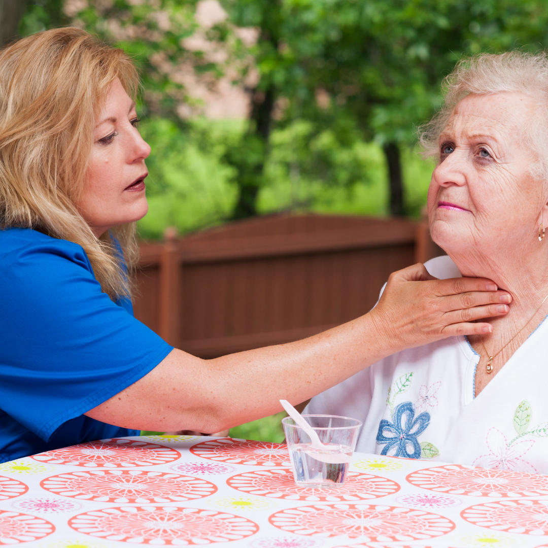 A therapist wearing a blue uniform gently touches an elderly woman's neck while she conducts a swallowing test.