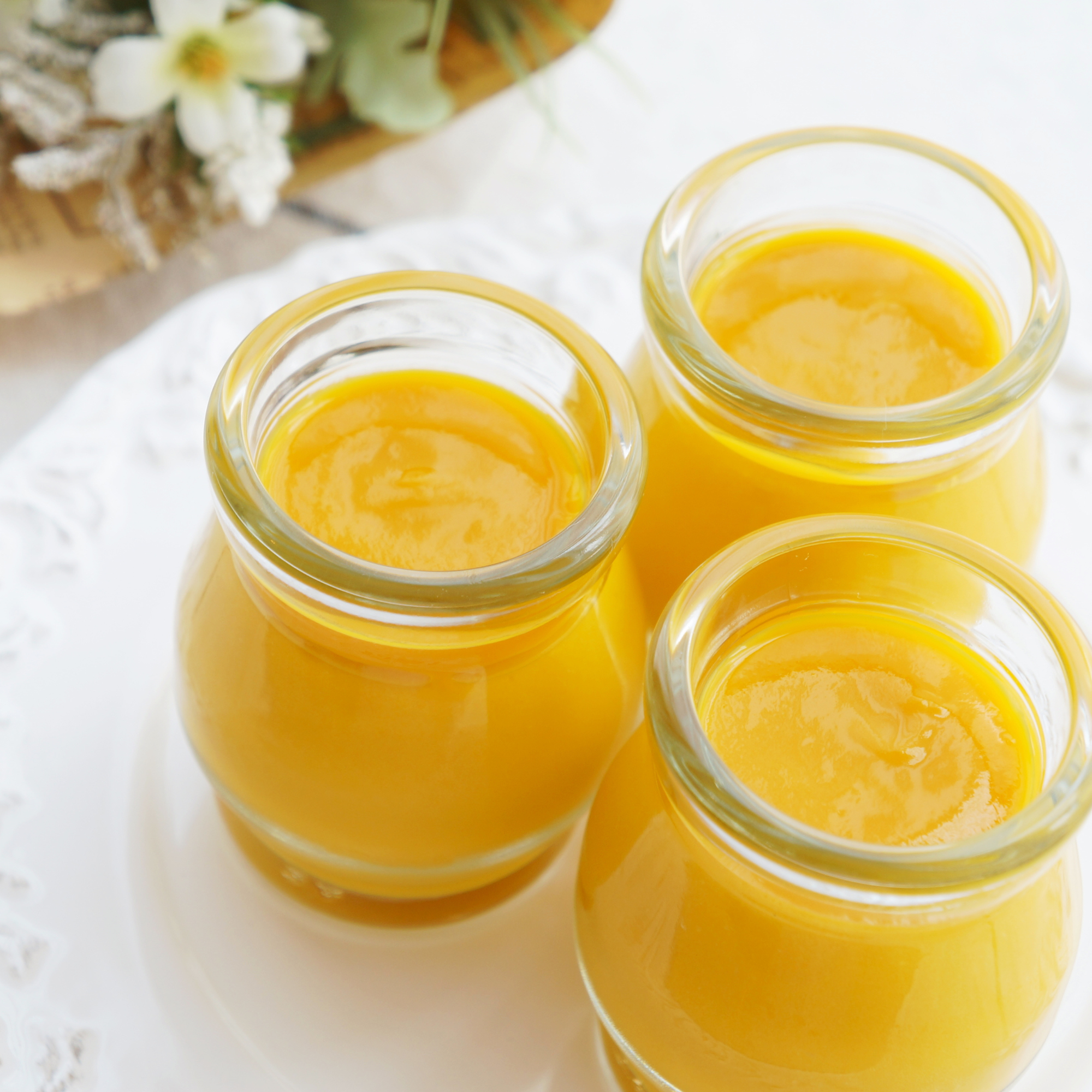 Two glass jars filled with yellow-orange liquid, possibly thickened juice, on a white surface with a blurred background of a small flower arrangement.