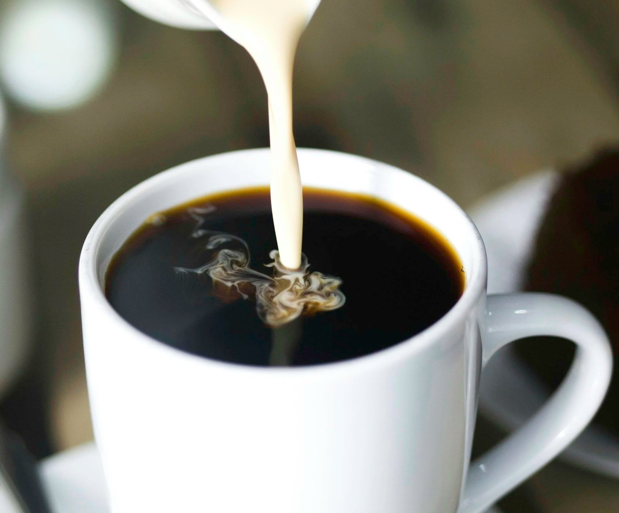 Close-up of a white ceramic cup filled with black coffee, with a stream of cream being poured into it.