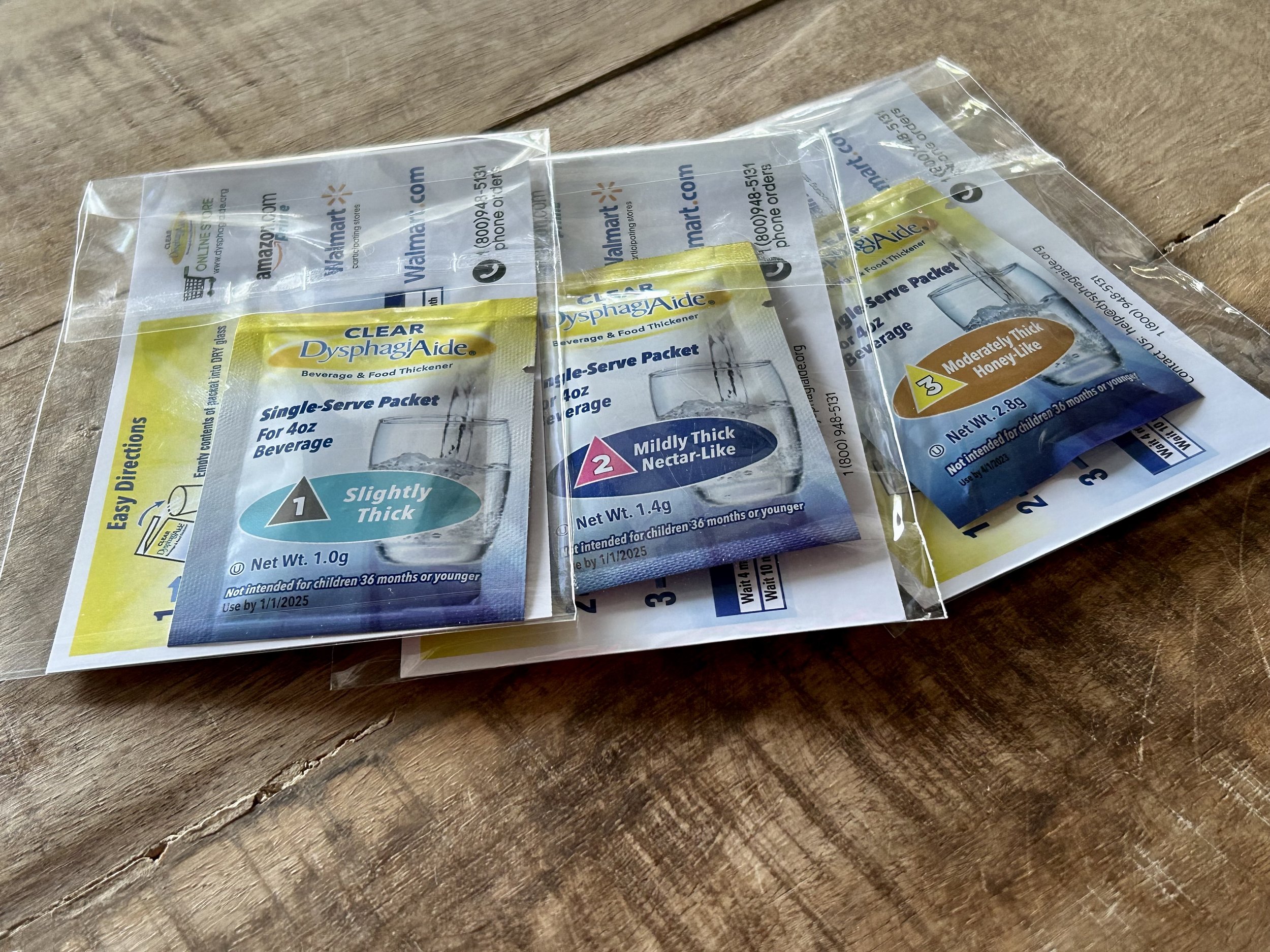 A clear plastic packet of DysphagiAide single-serve thickening powder for beverages, lying on a wooden surface.