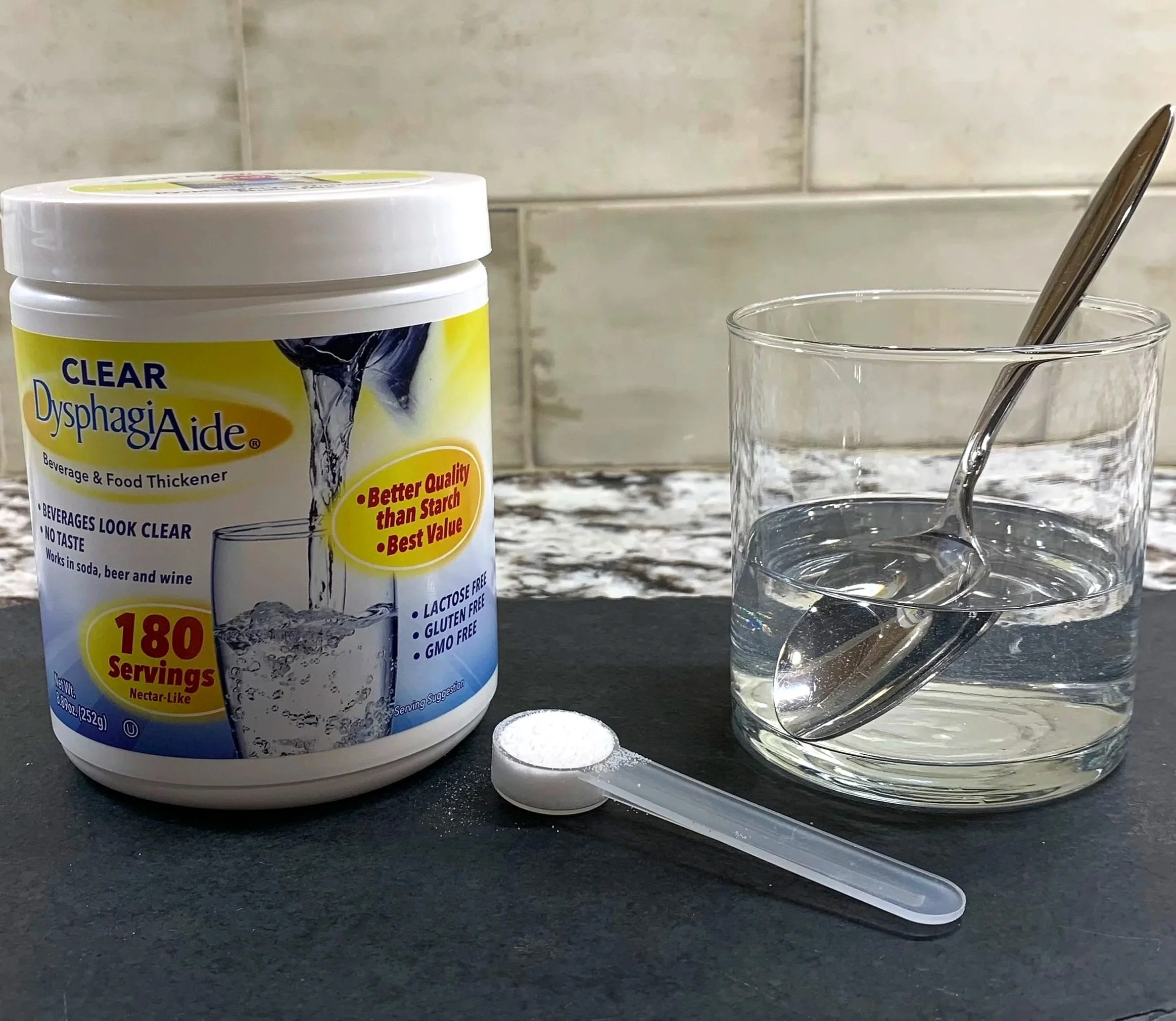 A container of Dysphagia Aide thickener, a glass of water, a metal spoon, and a white tablet on a black surface in front of a gray tiled wall.