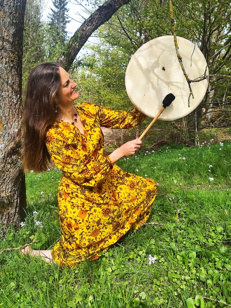 Eine lachende Frau in einem gelben, floral bedruckten Kleid, die im Wald auf den Knien sitzt und auf ein großes, rundes Trommeltuch schlägt.