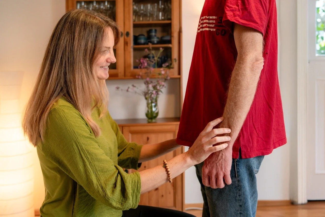 Eine Frau im grünen Hemd hält die Hand eines stehenden Mannes im roten T-Shirt, während sie lächelnd auf seinen Arm schaut.