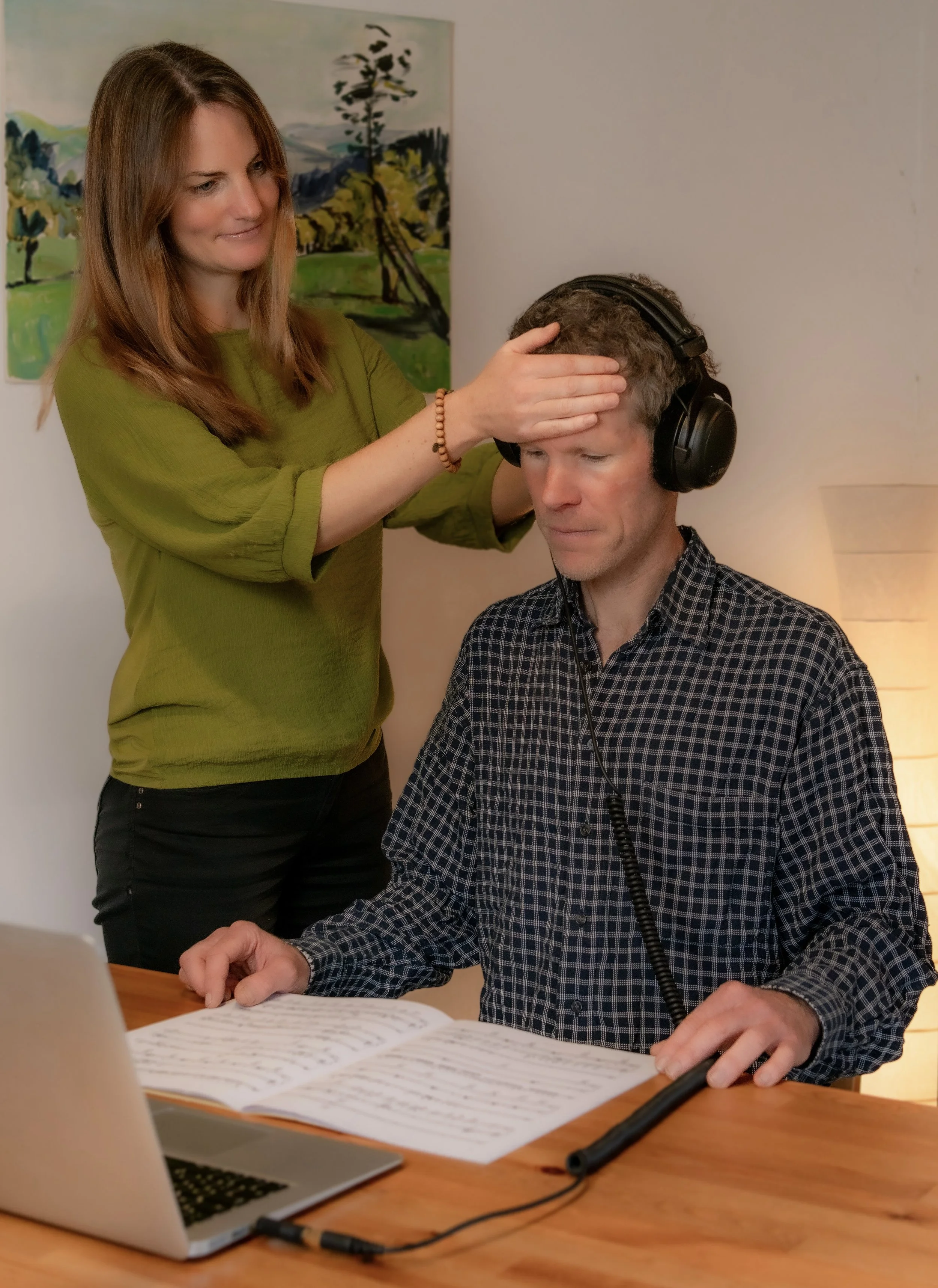 Eine Frau prüft die Stirn eines Mannes, der Kopfhörer trägt, während er ein Buch liest, während sie neben einem offenen Laptop steht.