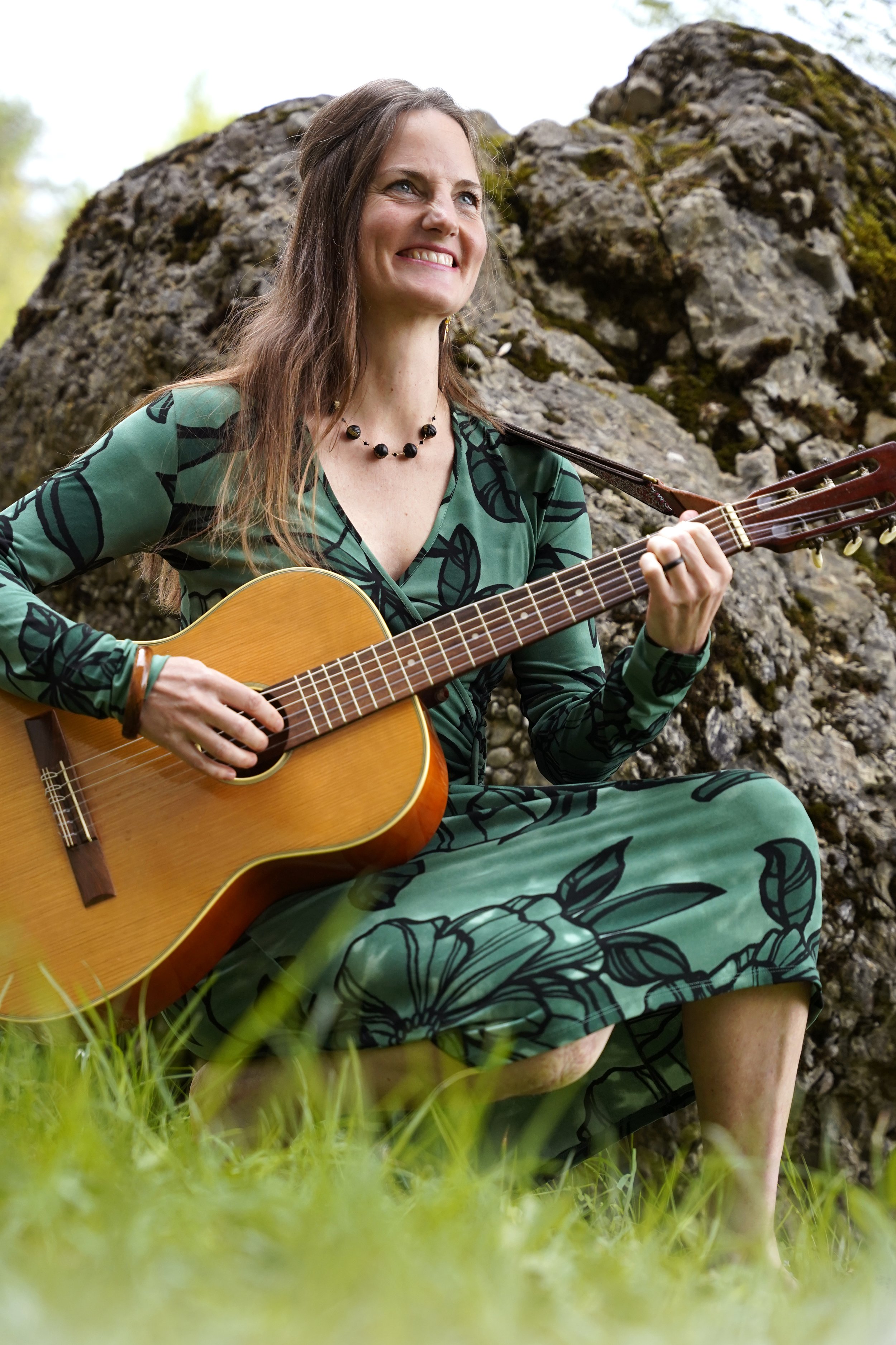 Frau spielt Gitarre im Freien vor Felsen, trägt grünes Kleid mit Blattmuster, sitzt auf Gras.