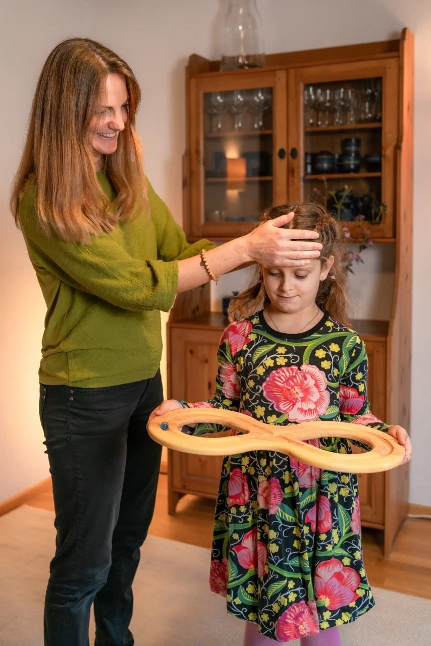 Eine Frau tätschelt das Mädchen auf dem Kopf, während sie ihm das Holzspielzeug zeigt. Das Mädchen schaut nach unten, in der Hand hält sie ein Holzspielzeug in Form einer acht. Im Hintergrund ein Holzschrank mit Gläsern und Geschirr.