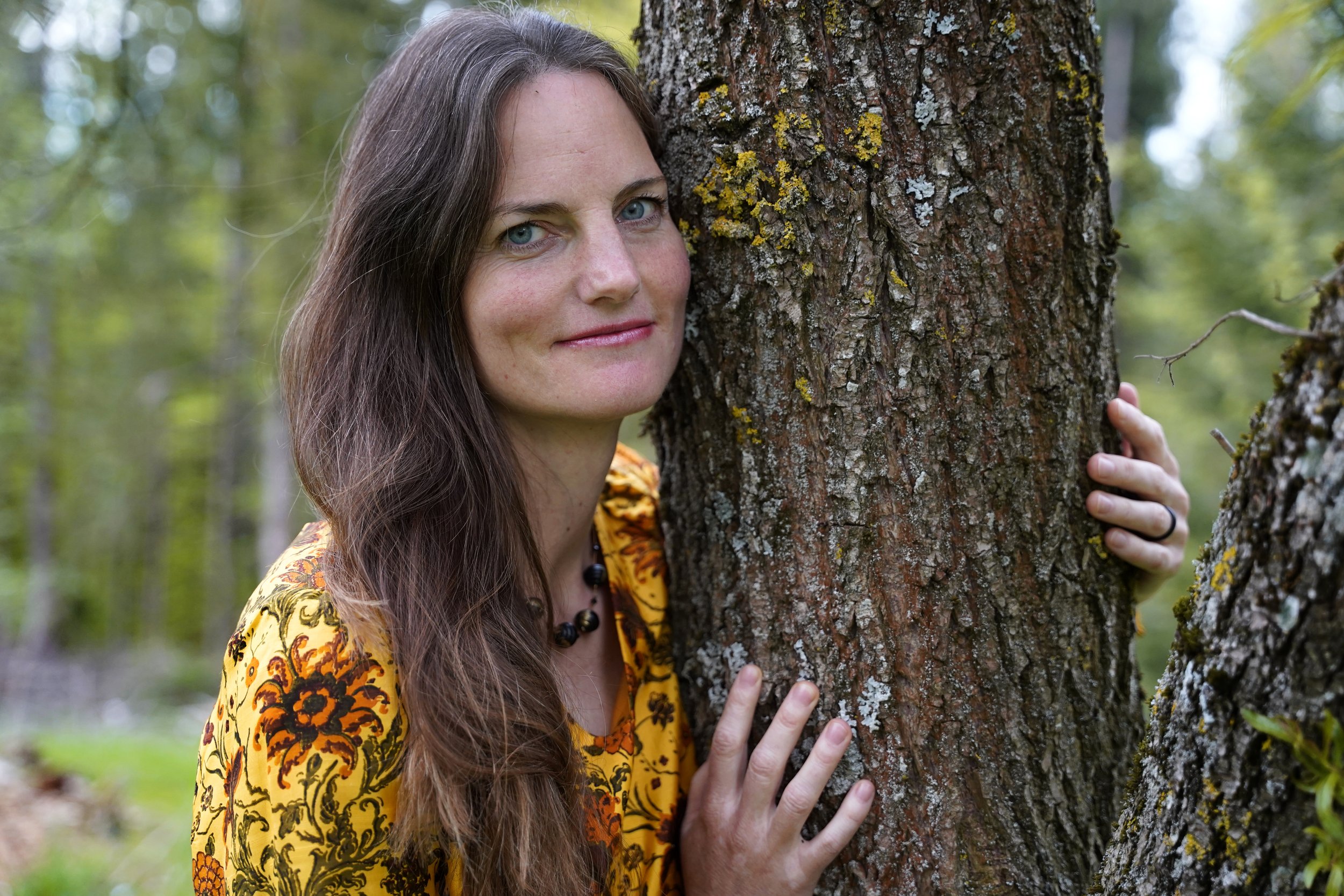 Eine Frau mit braunen Haaren und blauen Augen umarmt einen Baum im Wald, trägt ein gelbes Kleid mit orangefarbenen und schwarzen Mustern.