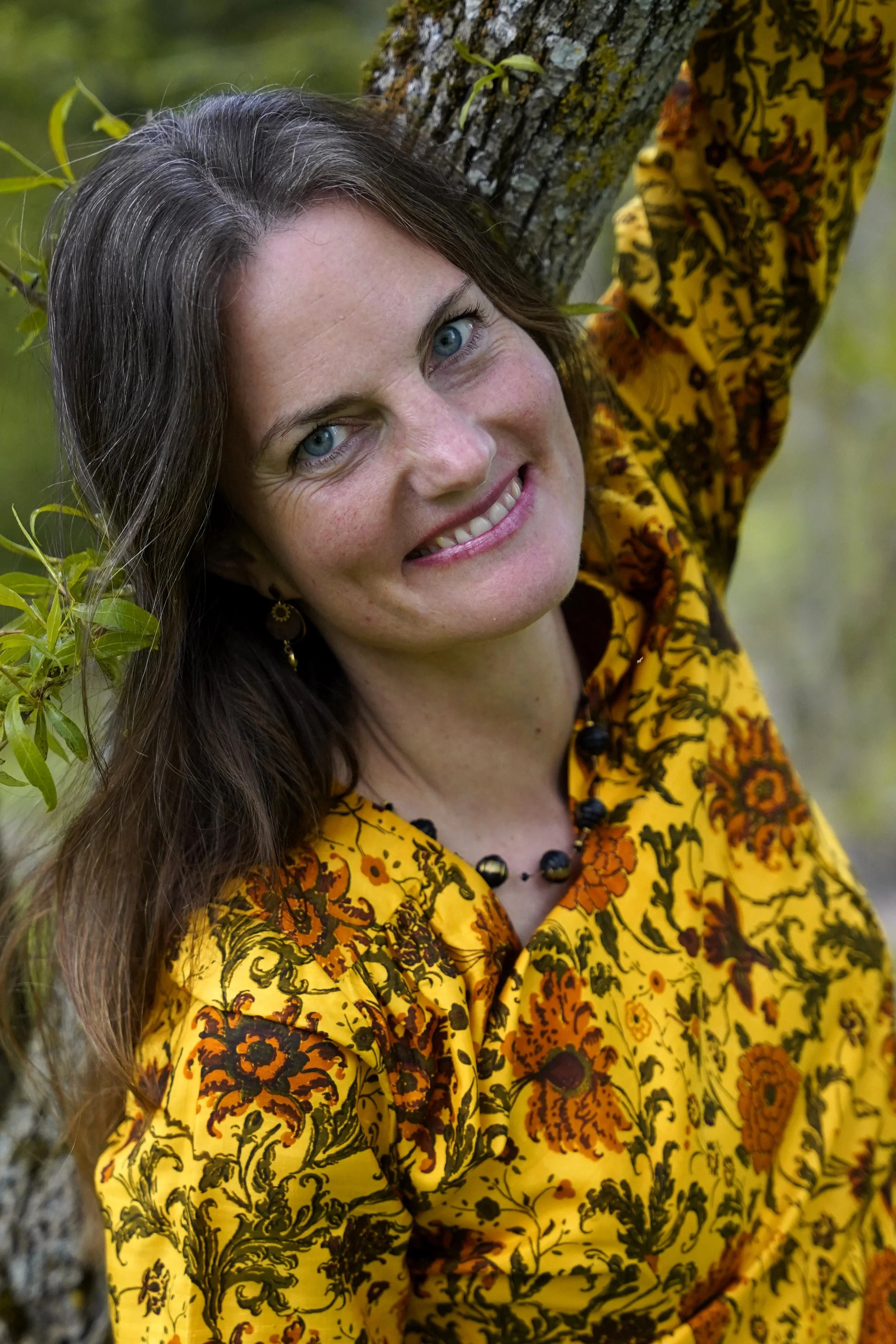 Nahaufnahme einer lachenden Frau mit blauen Augen, braunen Haaren und einem gelben Kleid mit schwarzen und orangefarbenen Mustern, vor einem Baum mit grünen Blättern im Hintergrund.