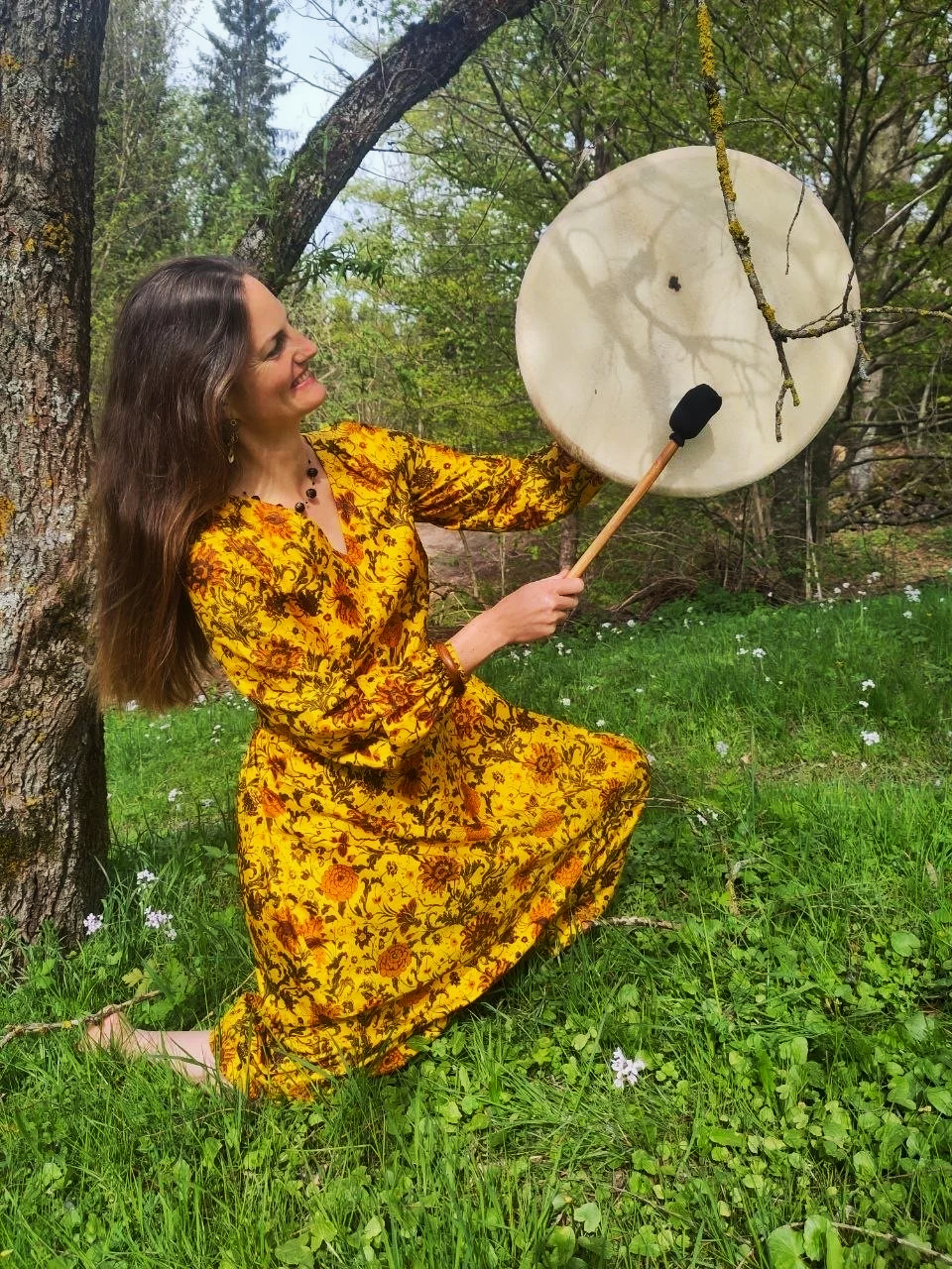 Frau in gelbem, floral gemustertem Kleid sitzt im Grünen neben einem Baum, hält eine große weiße Trommel und lächelt in die Kamera.