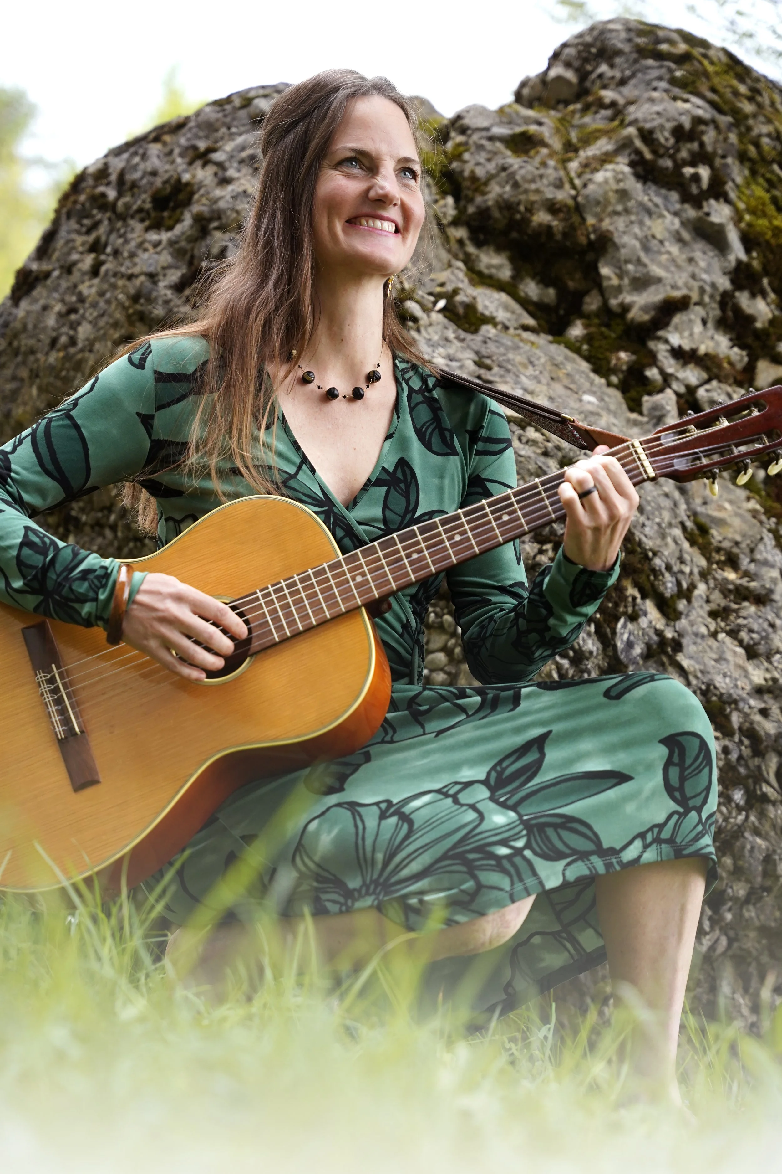 Frau sitzt im Freien auf einem Felsen, spielt Gitarre und lächelt, umgeben von grüner Natur.