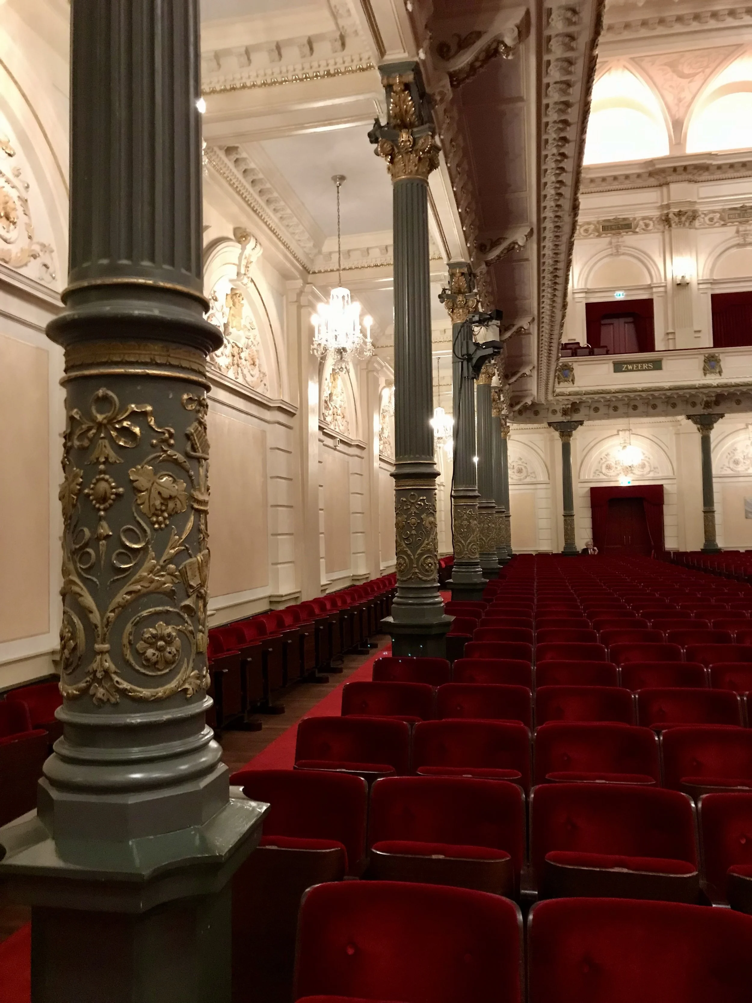 Ein beeindruckender Innenraum eines historischen Theaters oder Opernhauses mit roten Sitzreihen, kunstvollen Dekorationen, goldverzierten Säulen und großen Kristallleuchtern.
