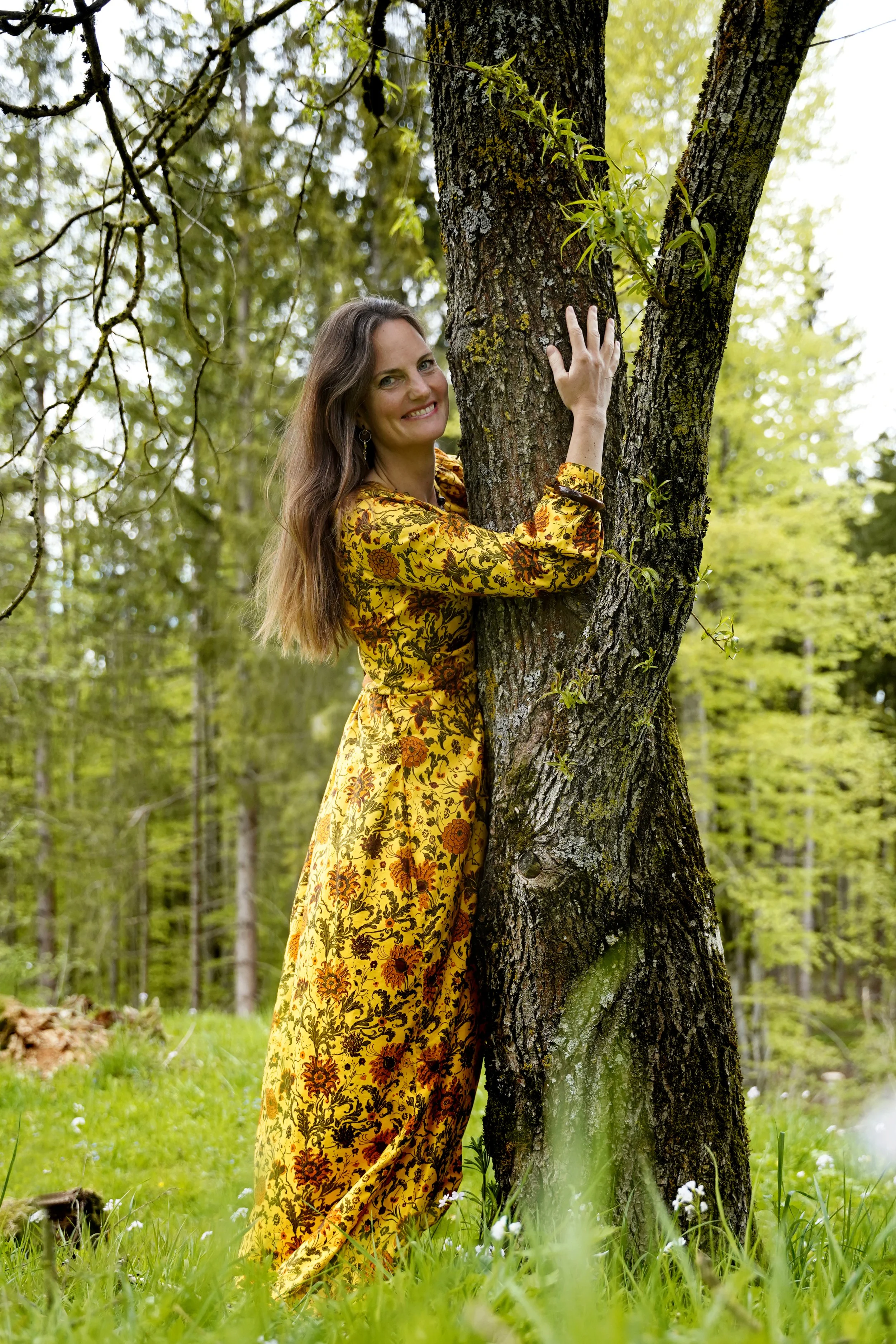 Eine Frau in einem gelben, blumenmusternden Kleid umarmt einen Baum in einem grünen Wald. Ihr Lächeln ist sichtbar, sie hat braune, lange Haare und sieht glücklich aus.