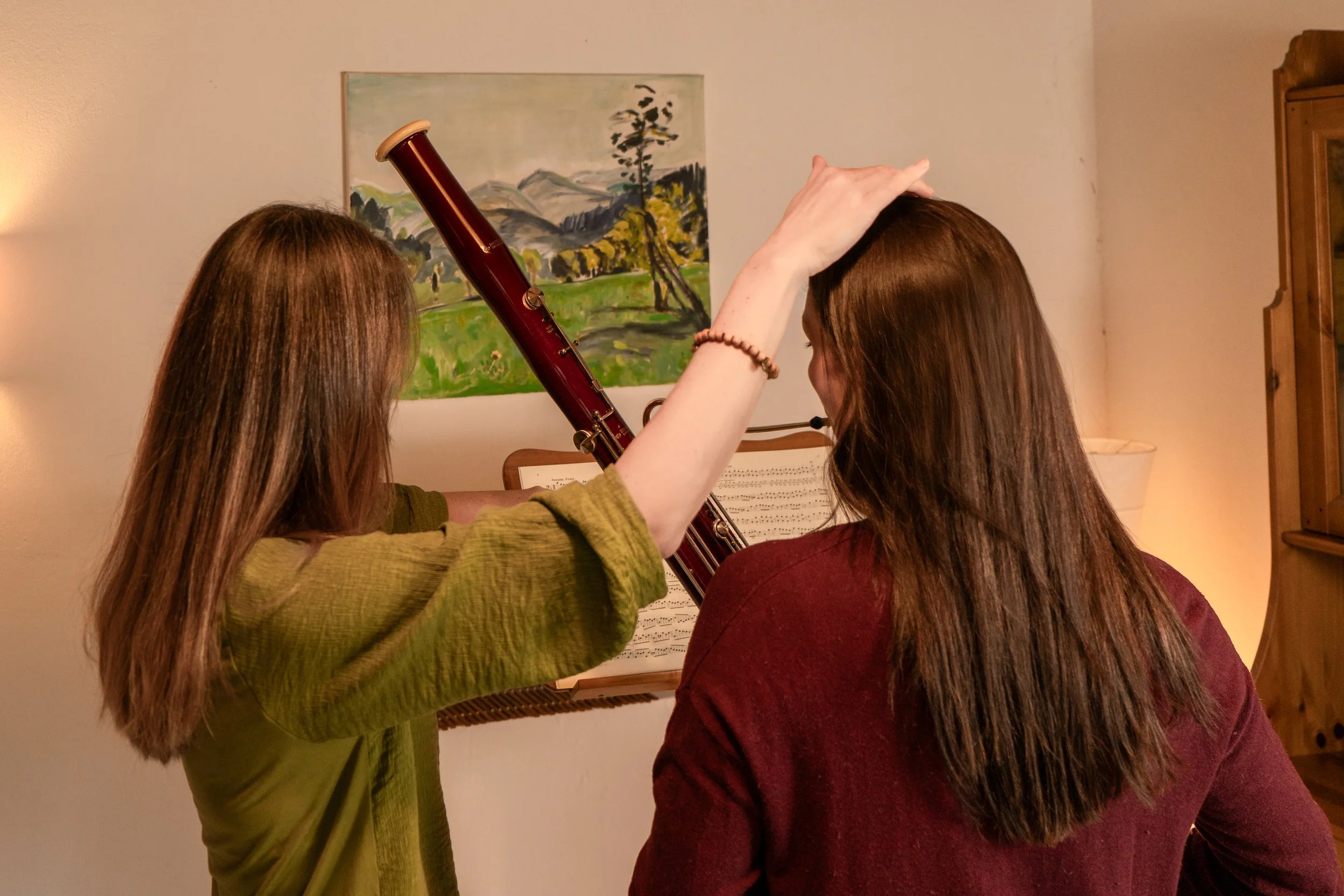 Zwei Frauen stehen vor einem Notenblatt und einem Musikinstrument, während sie auf ein Klavier schauen, in einem Raum mit einem Wandbild und einem Holzschrank.
